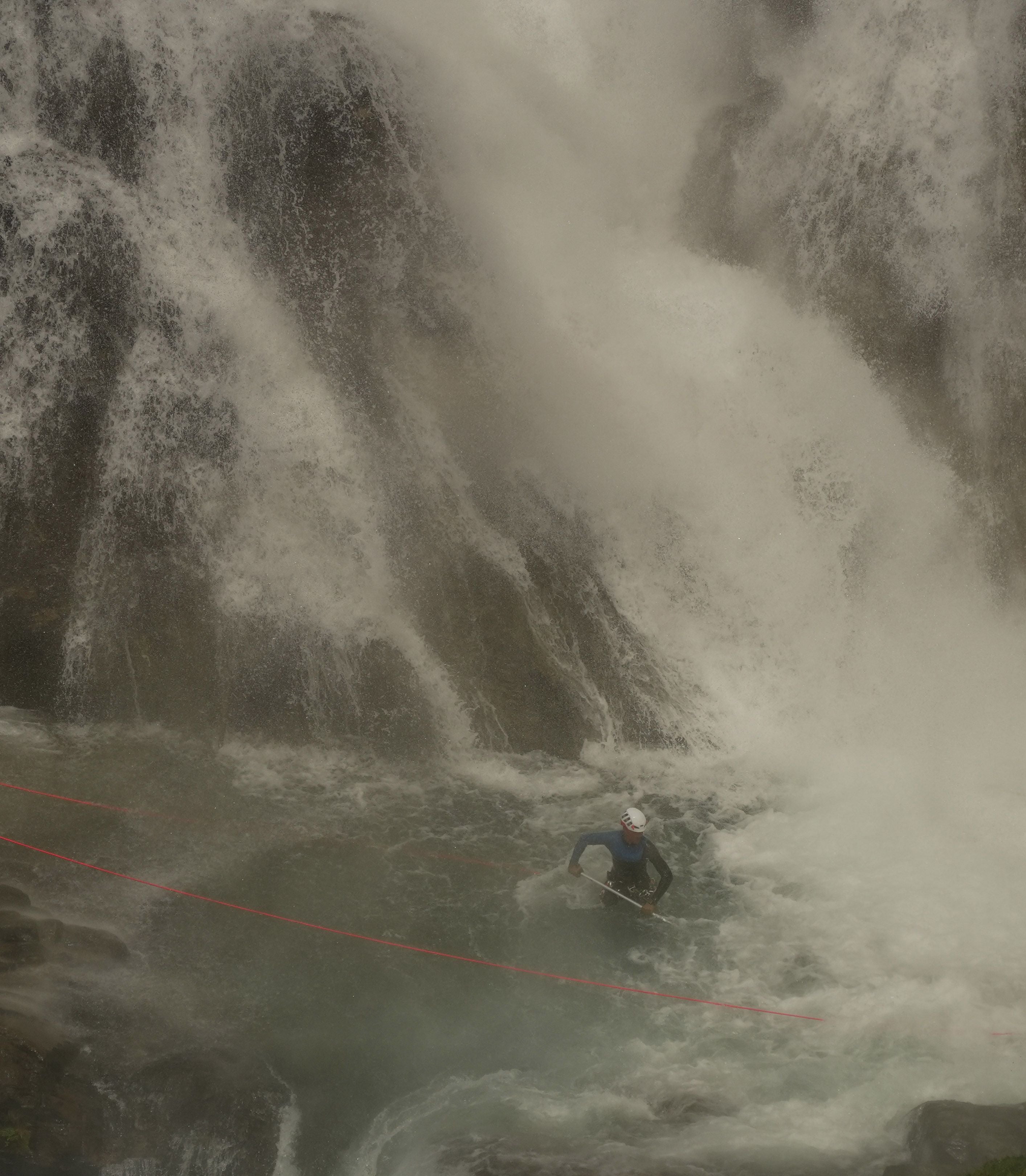 Person mit Helm watet an einem Wasserfall durch das Wasser, gesichert mit einem roten Seil.