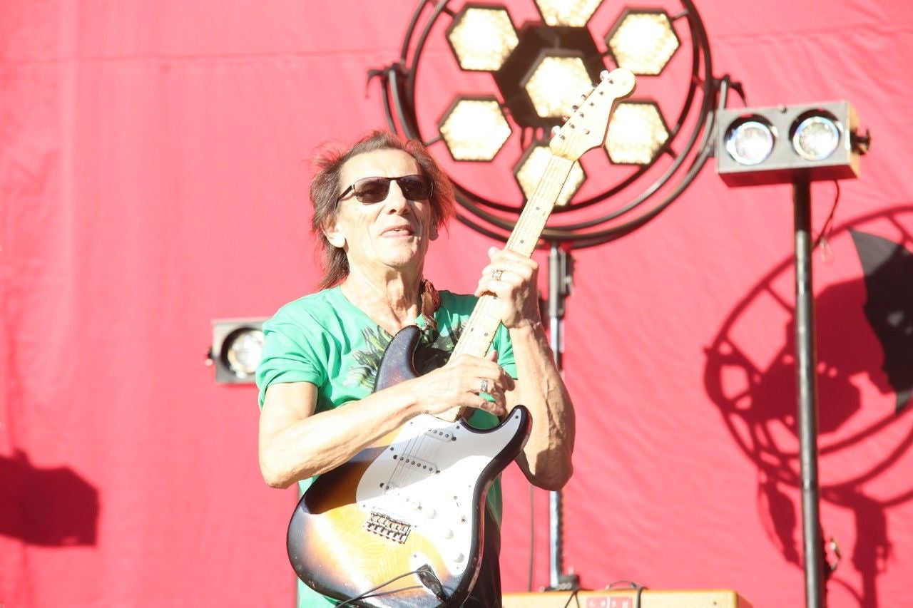 Ein Mann mit Sonnenbrille spielt E-Gitarre vor rotem Hintergrund und Scheinwerfern.
