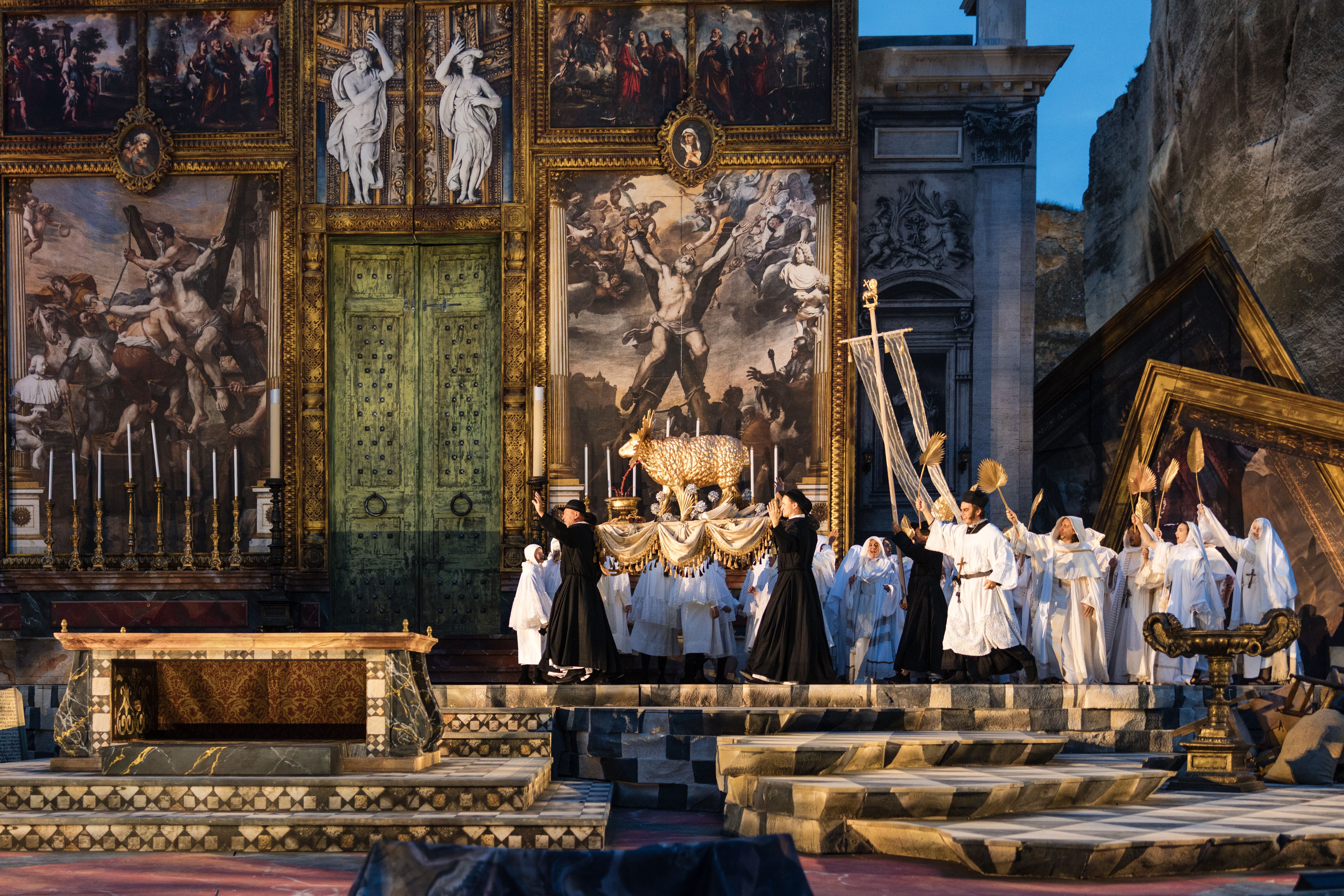 Bühnenszene aus der Oper "Tosca" mit vielen Darstellern in weißen Gewändern und prächtigem Bühnenbild.