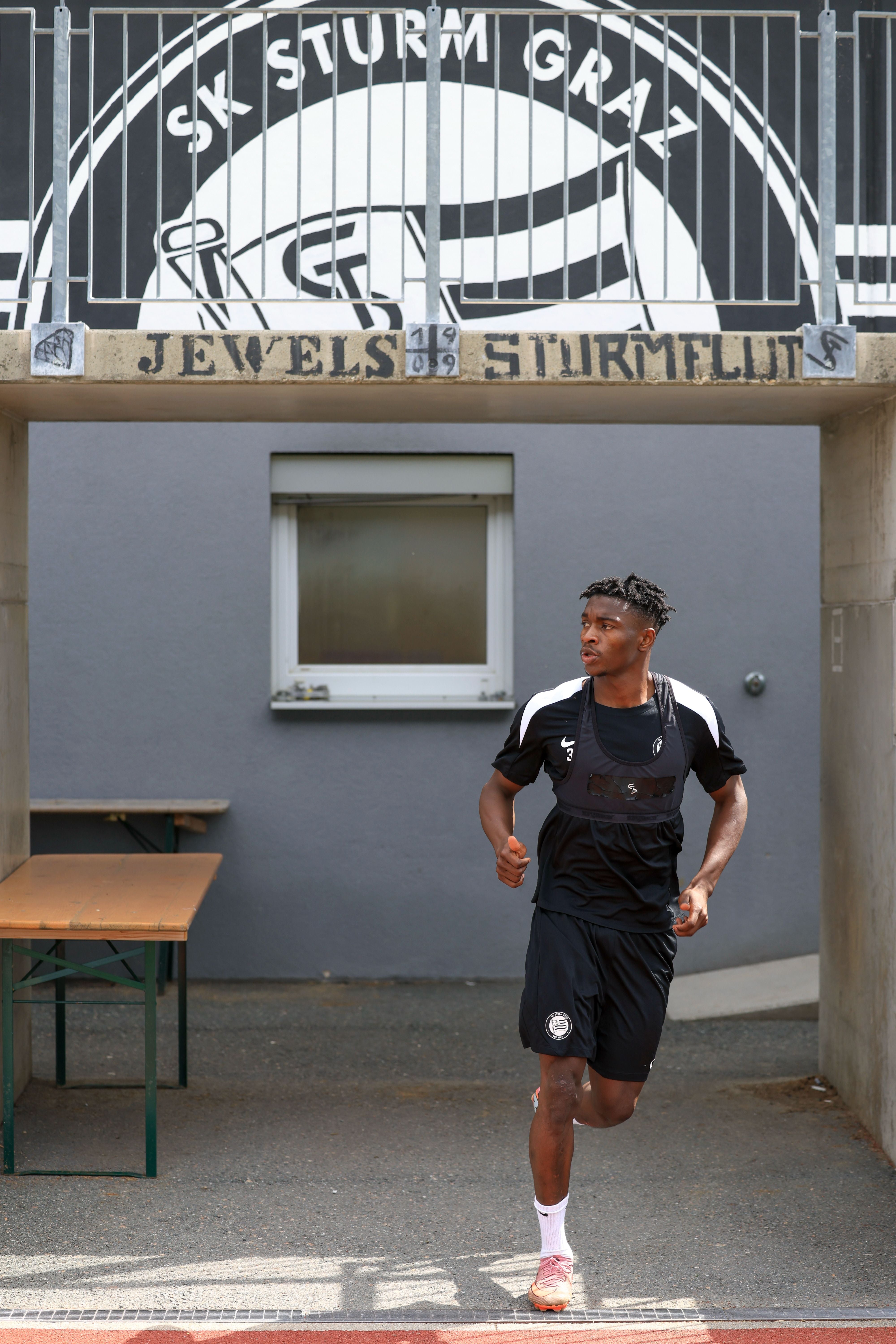 Ein Fußballspieler von Sturm Graz läuft im Trainingsanzug unter einem Vereinsbanner hindurch.