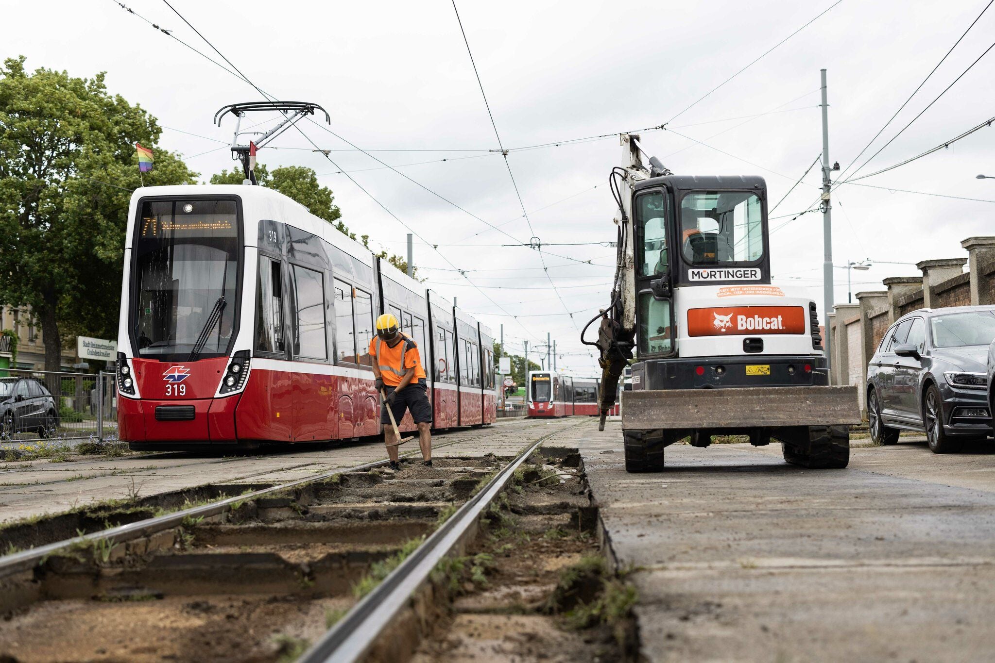 Bauarbeiten an Straßenbahngleisen mit Arbeiter, Bagger und einer roten Straßenbahn.