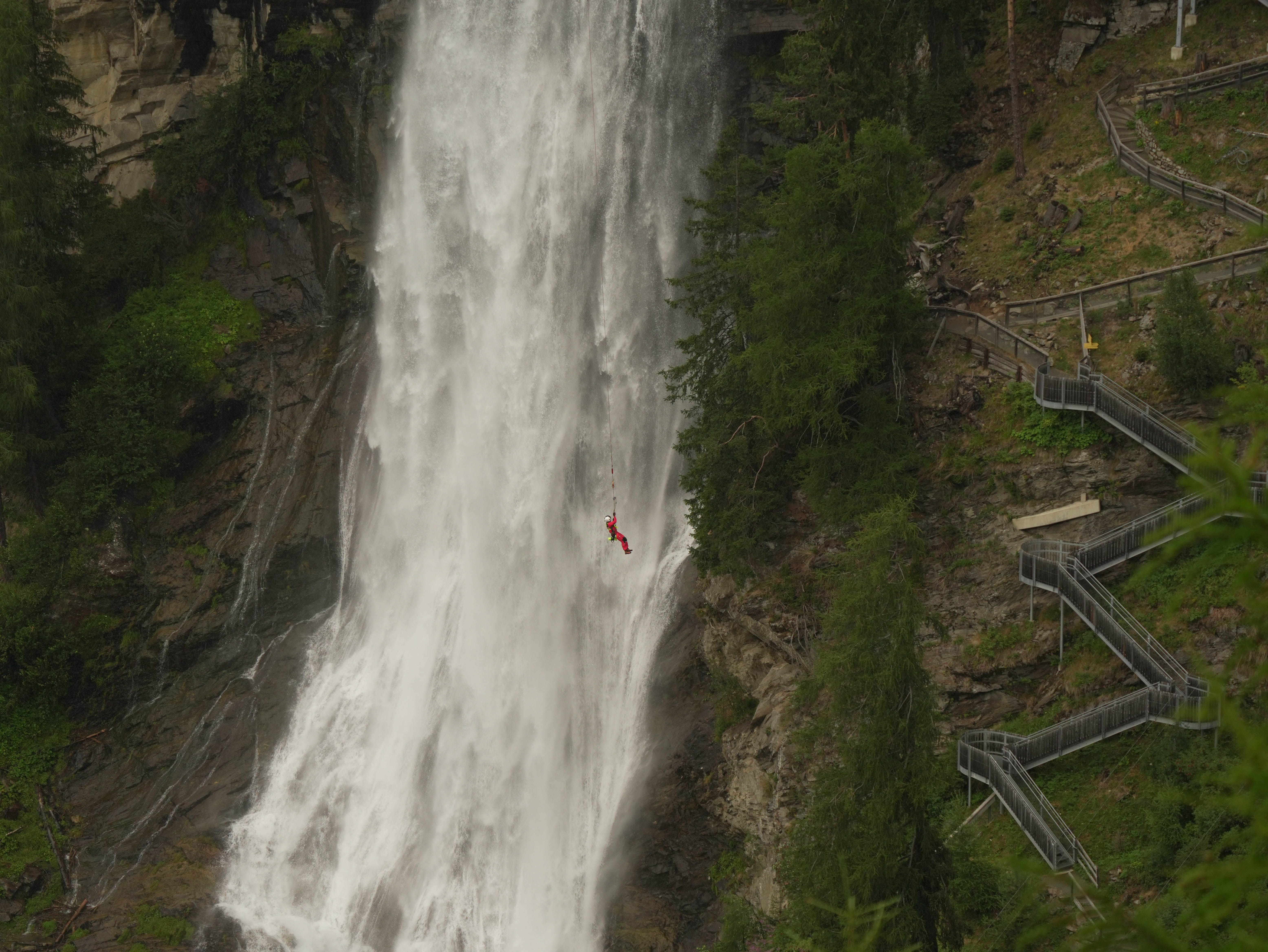 Person beim Abseilen mitten durch einen großen Wasserfall mit steilem Wanderweg rechts.