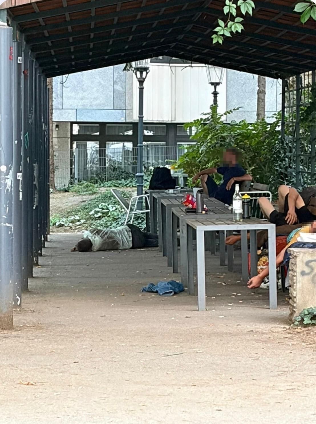 Mehrere Menschen sitzen oder liegen unter einem überdachten Platz im Freien, teils auf dem Boden.