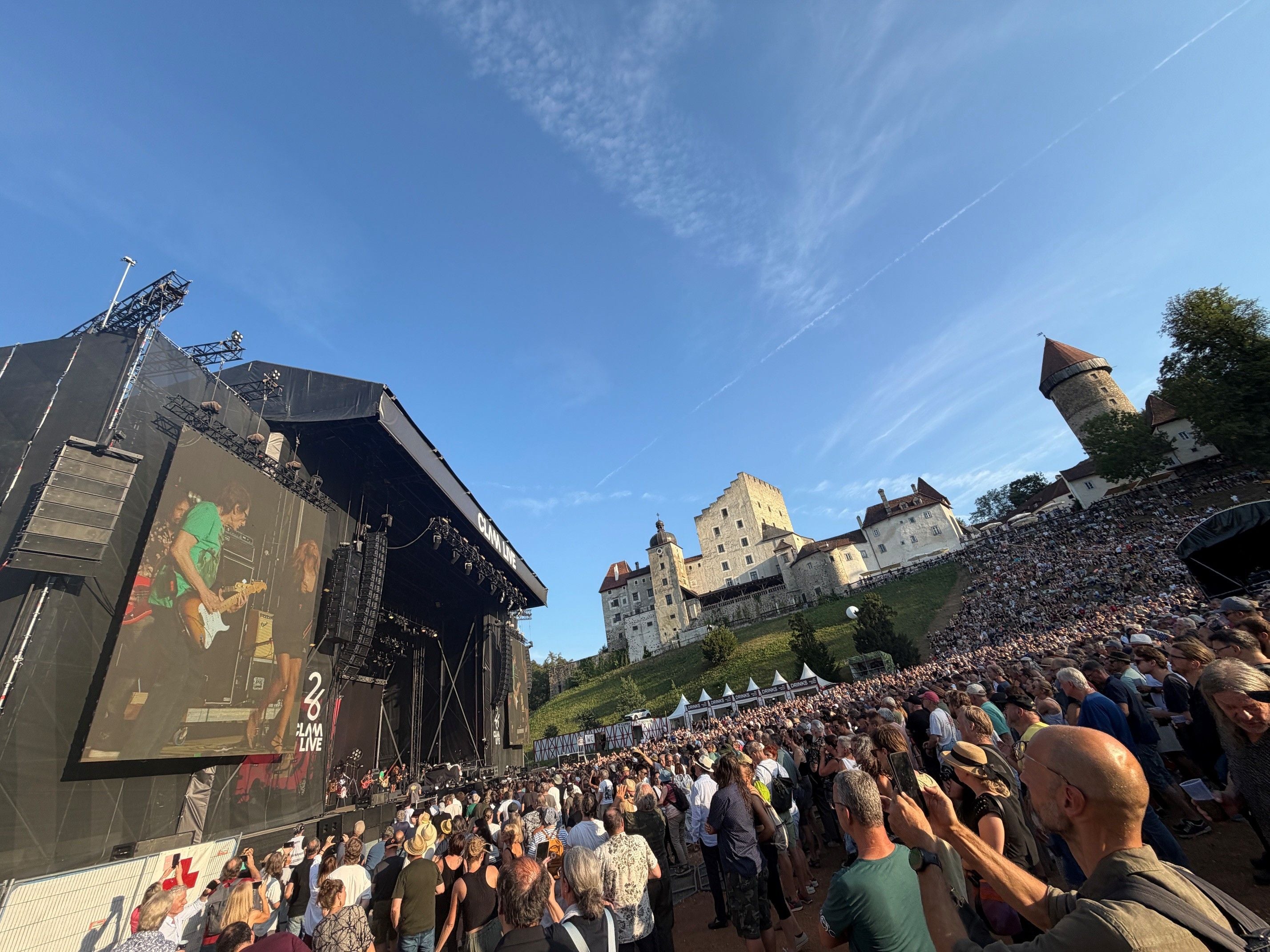 Open-Air-Konzert vor einer großen Menschenmenge mit Bühne und Burg im Hintergrund bei Sonnenschein.