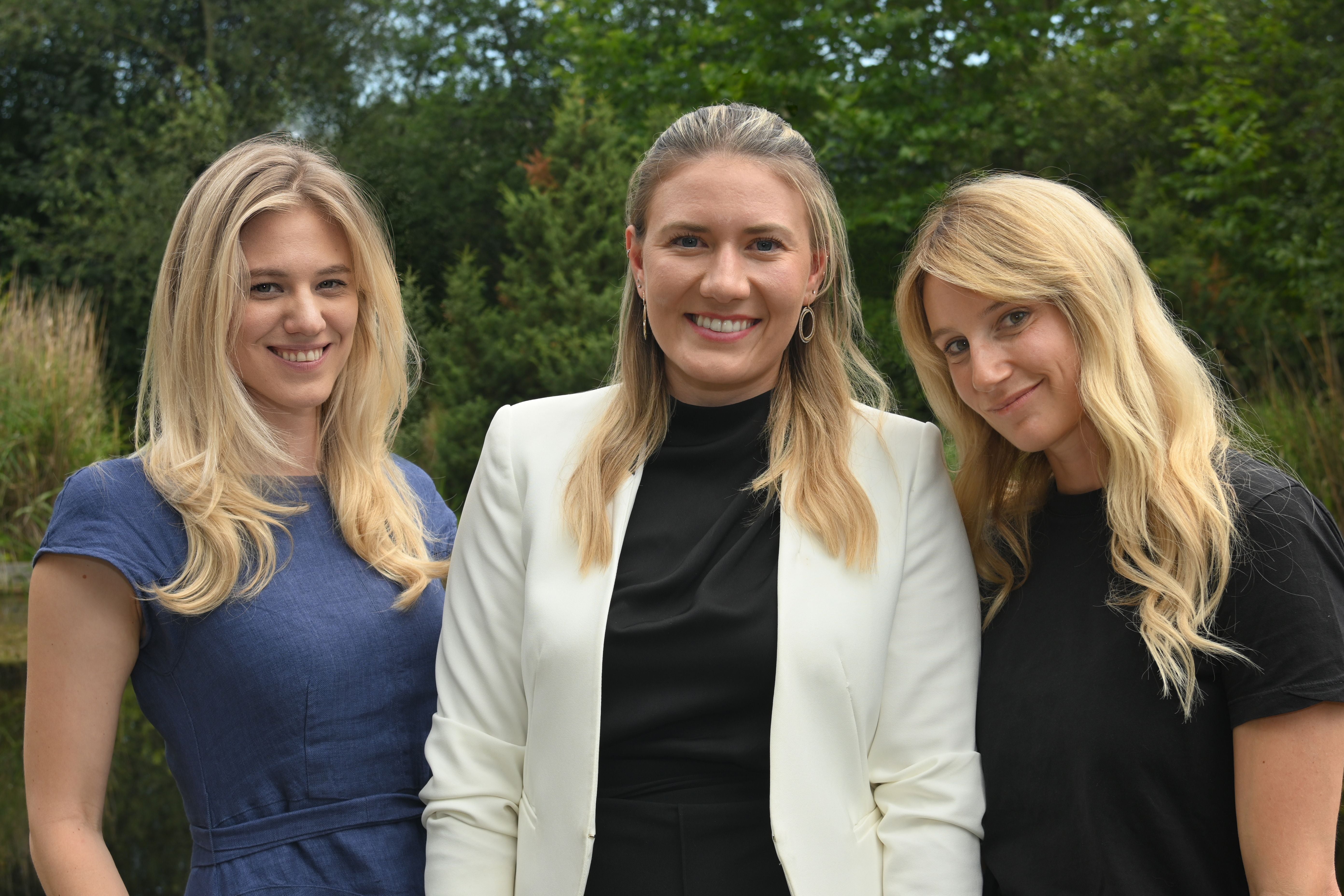 Drei Frauen mit langen blonden Haaren posieren lächelnd im Freien vor grünen Bäumen.