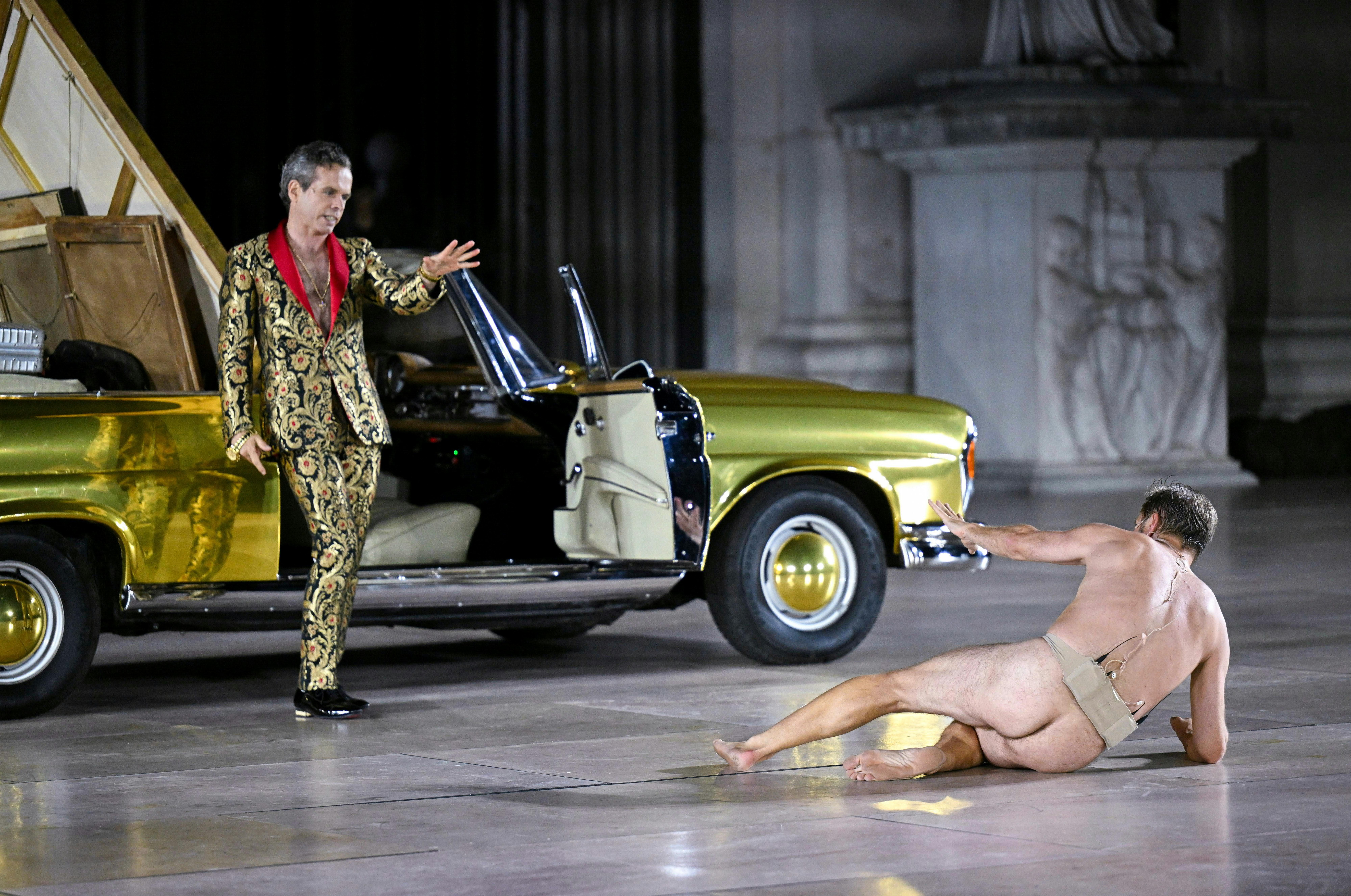 Zwei Schauspieler spielen vor einem goldenen Auto auf einer Bühne, einer steht, einer liegt am Boden.