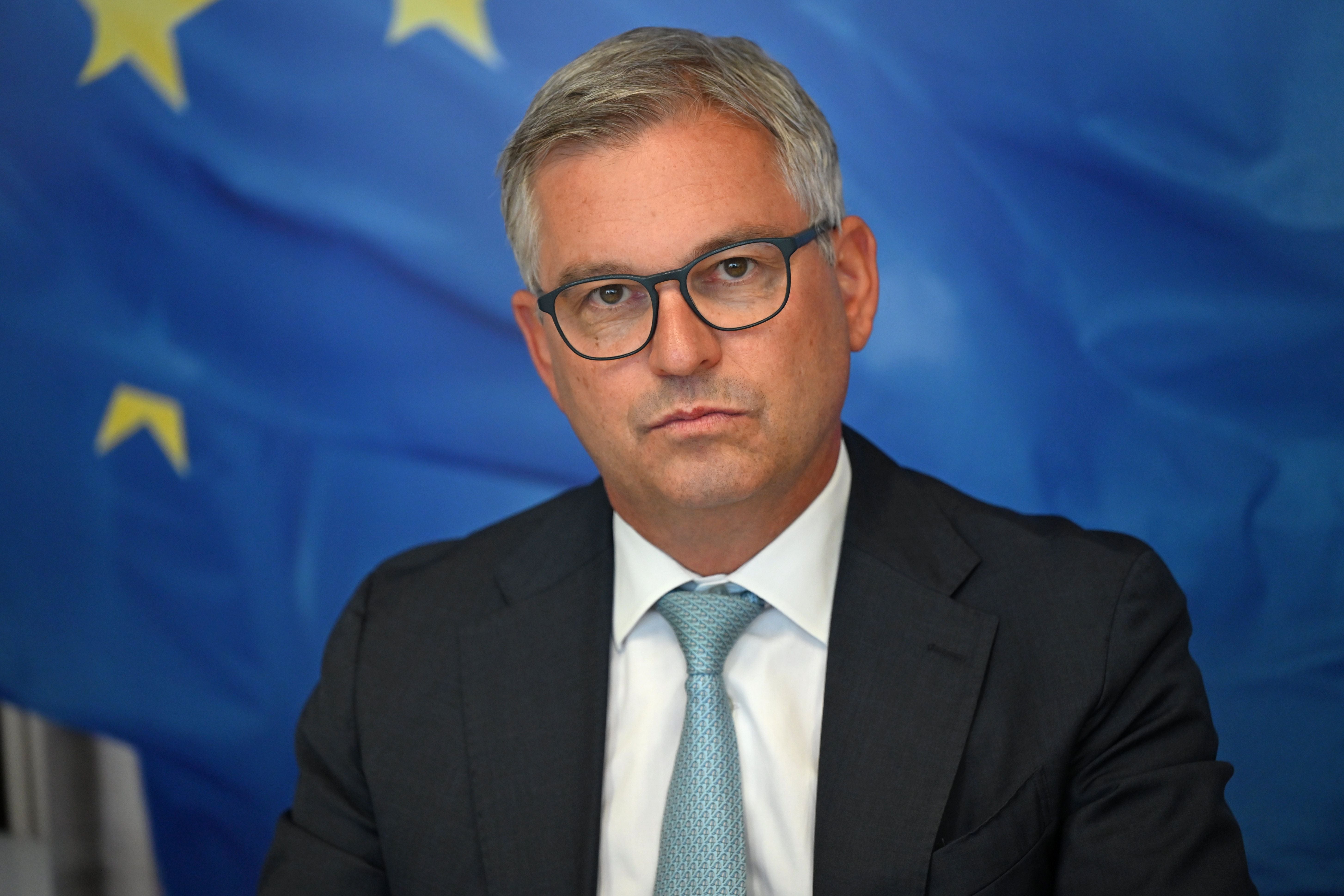 Ein Mann im Anzug vor einer blauen Flagge mit gelben Sternen, die EU-Flagge darstellend.