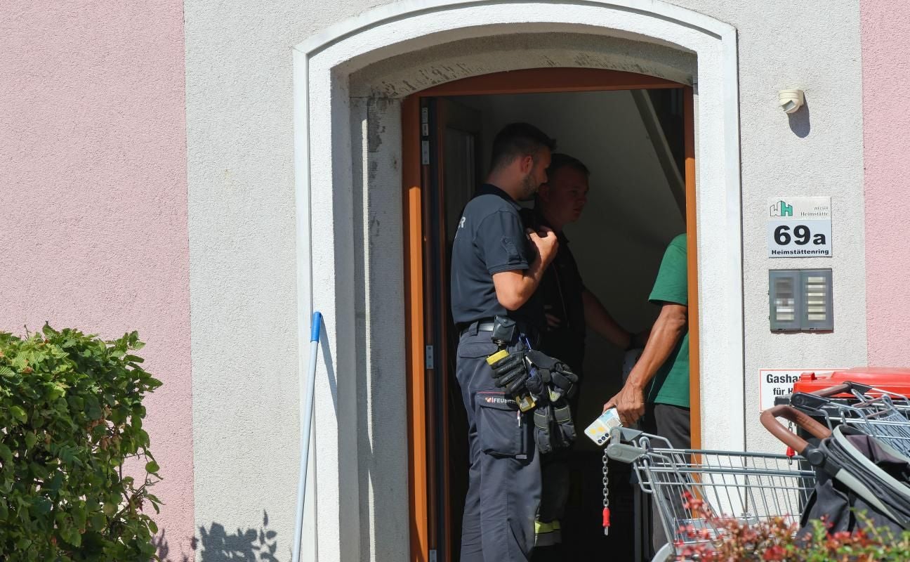 Zwei Feuerwehrleute und ein Mann stehen im Eingangsbereich eines Wohnhauses.