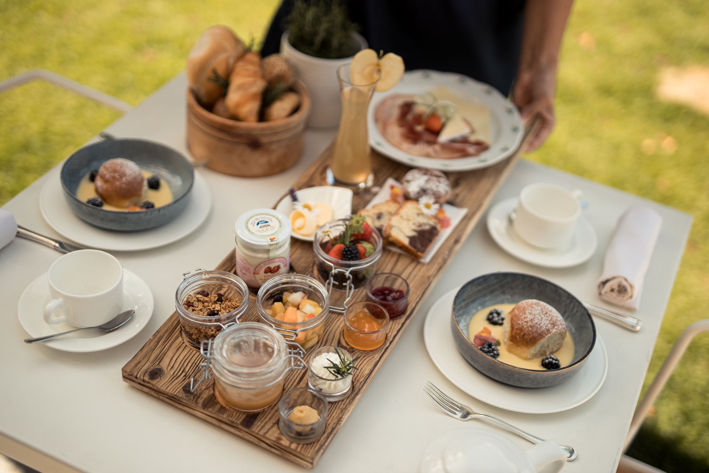Reich gedeckter Frühstückstisch mit Gebäck, Obst, Joghurt, Saft und Süßspeisen im Freien.