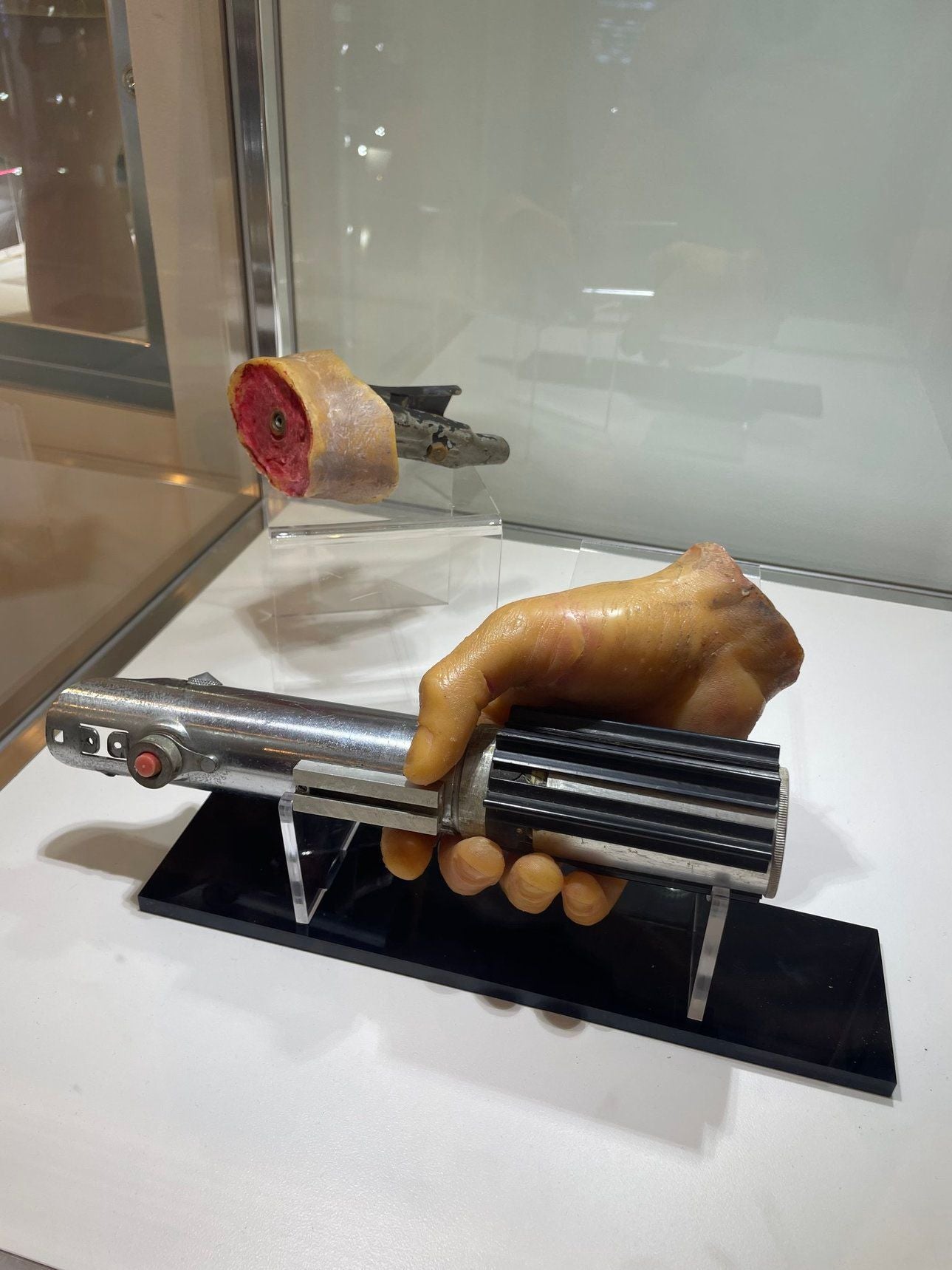 Künstliche Hand hält ein Lichtschwert, dahinter liegt eine abgeschnittene Hand in einer Vitrine.