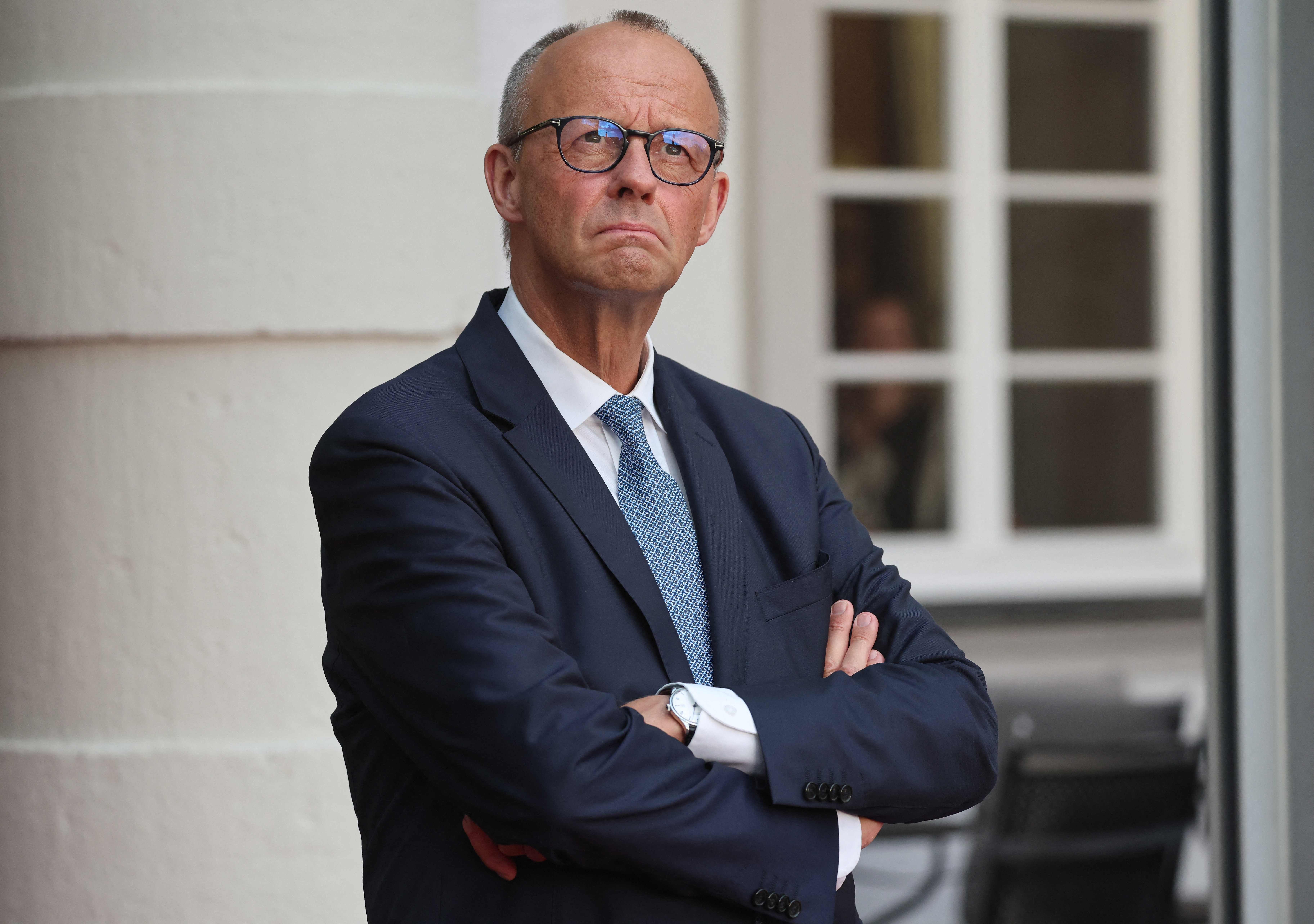 Ein Mann im Anzug steht mit verschränkten Armen vor einem Gebäude und blickt ernst.