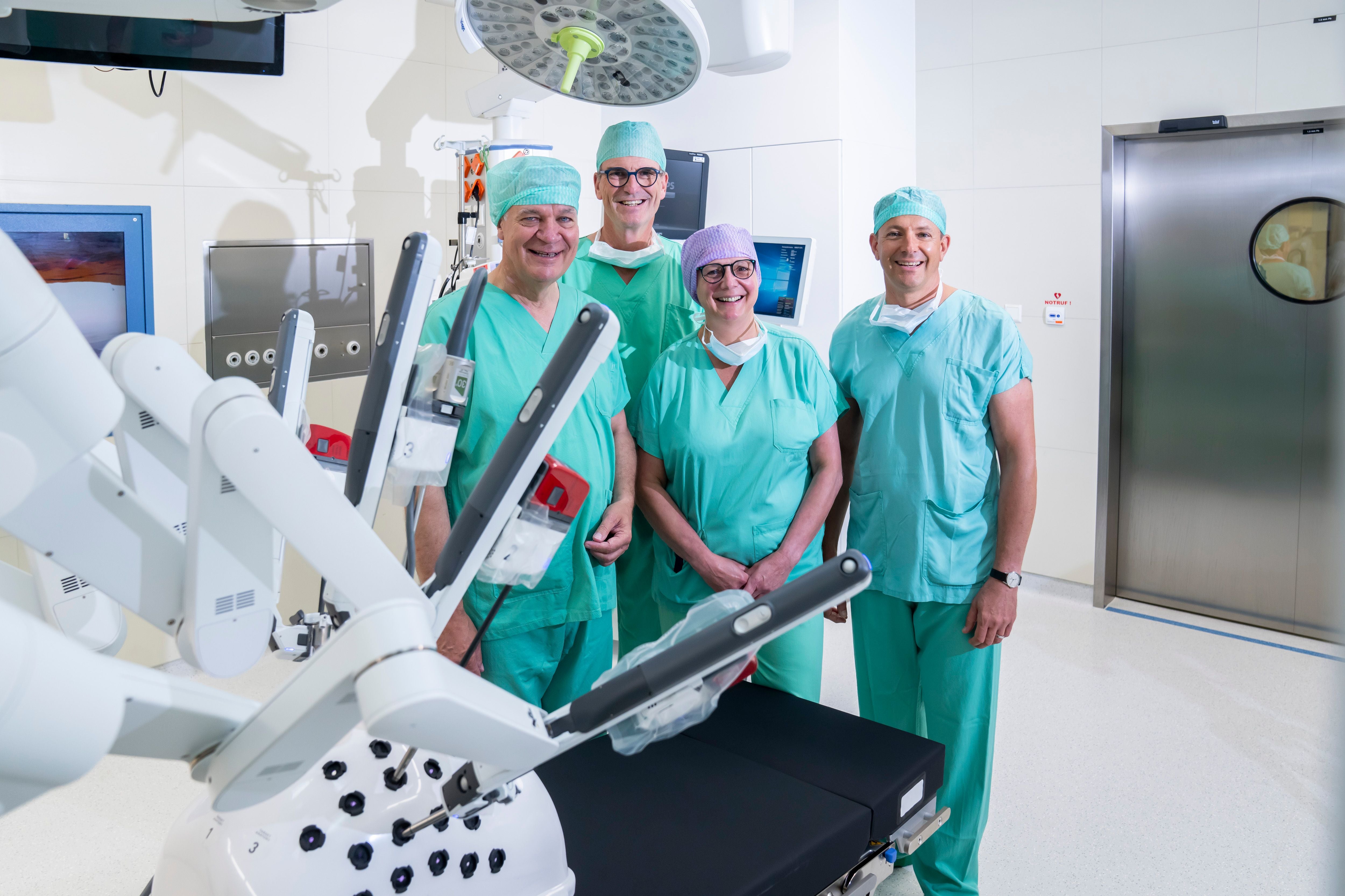 Vier Chirurgen stehen lächelnd neben einem OP-Roboter in einem modernen Operationssaal.