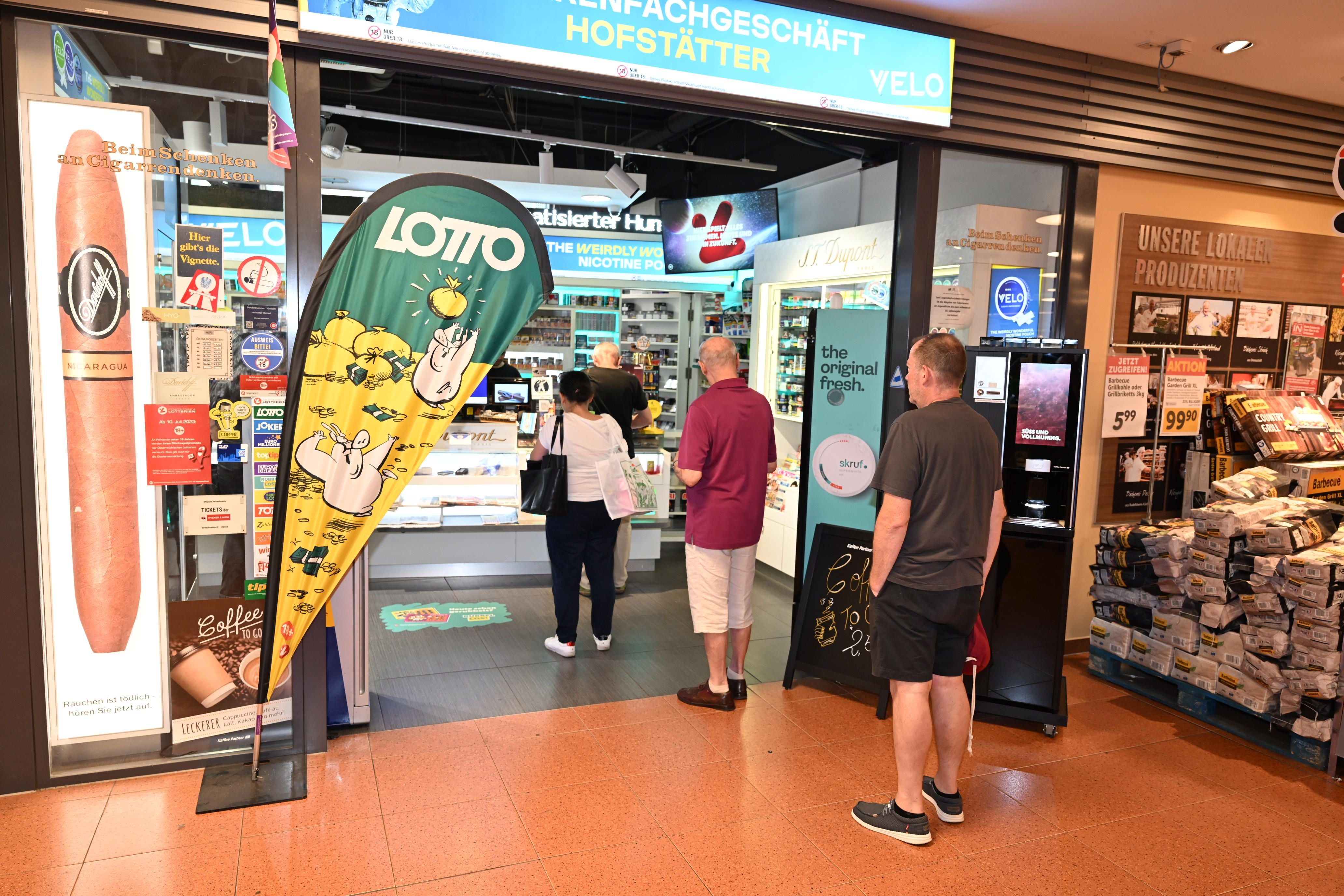 Mehrere Menschen stehen in einer Schlange vor einem Lotto- und Tabakgeschäft im Einkaufszentrum.