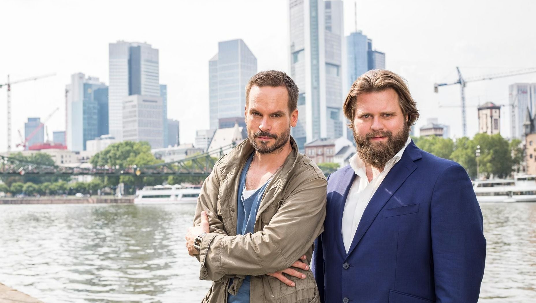 Zwei Männer stehen am Flussufer vor moderner Skyline in Frankfurt am Main.