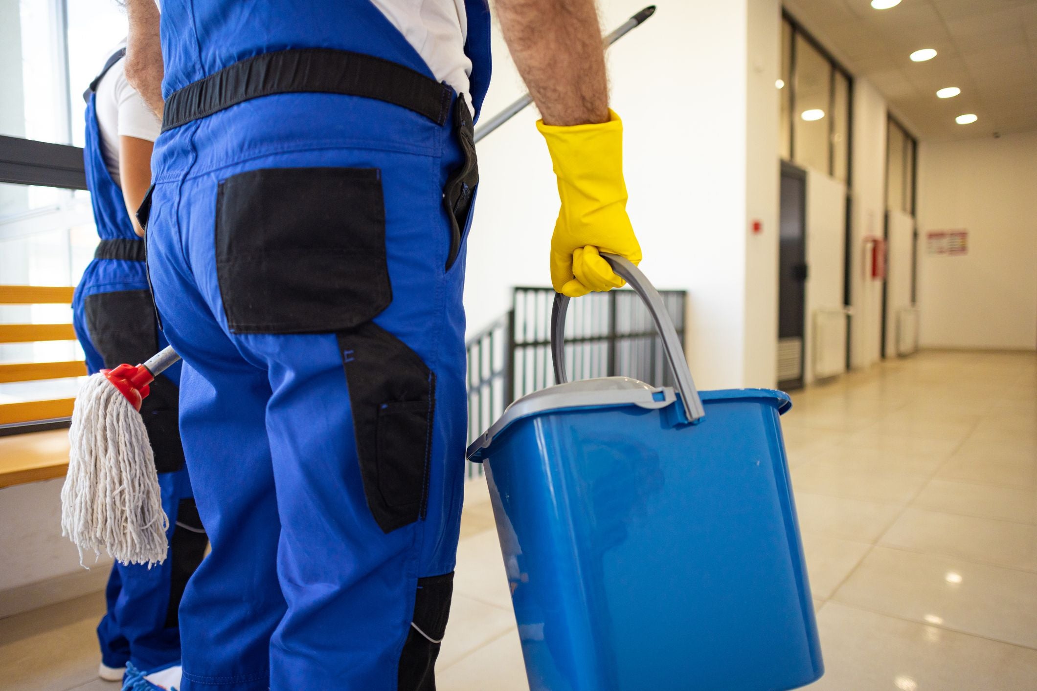 Zwei Reinigungskräfte tragen Putzutensilien wie Eimer und Wischmopp in einem Flur.