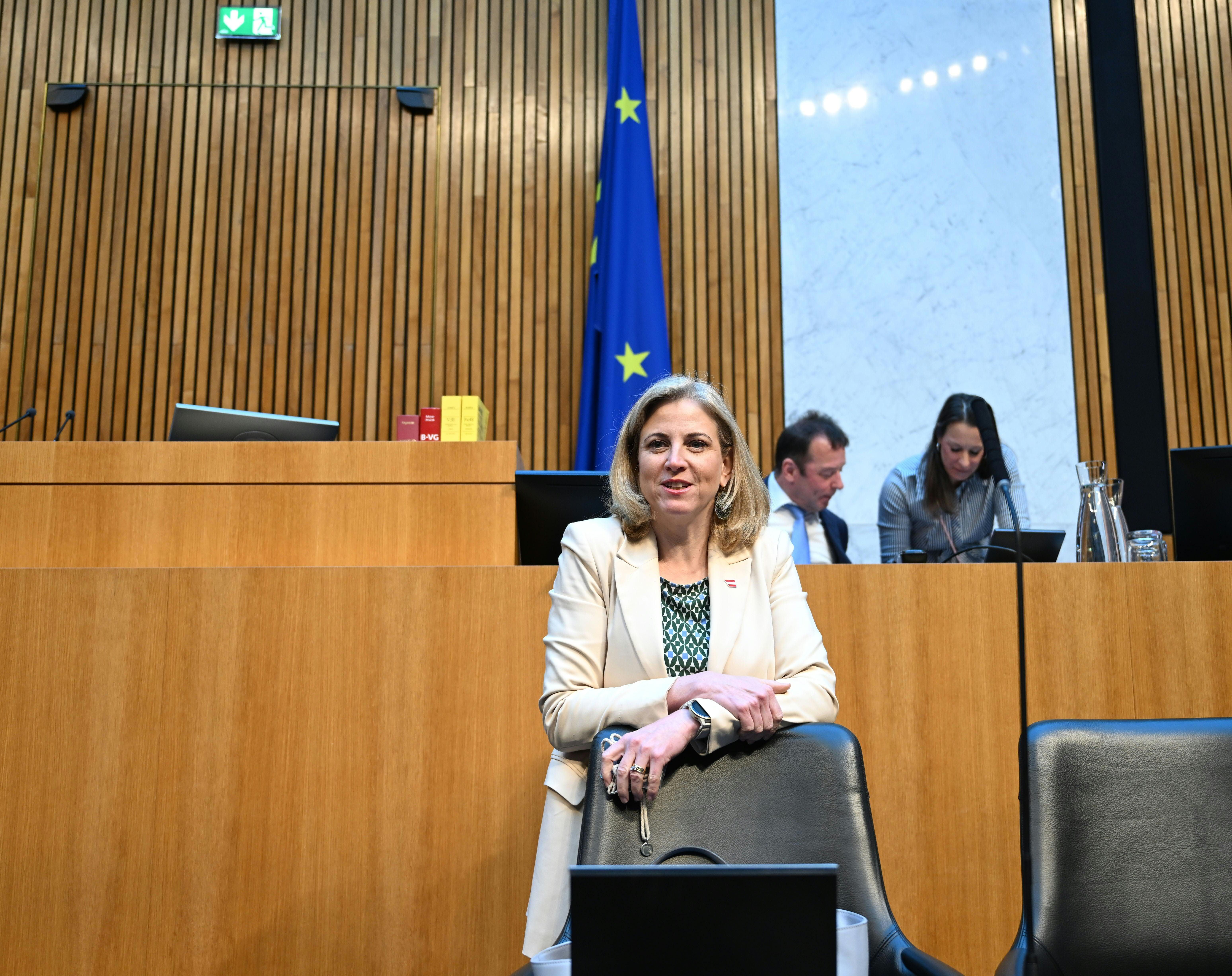 Eine Frau steht im Parlament vor leeren Sitzen, im Hintergrund arbeiten zwei Personen am Computer.