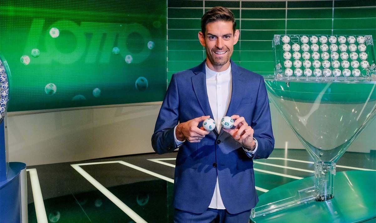 Ein Mann im Anzug hält zwei Lotto-Kugeln in einem Fernsehstudio.