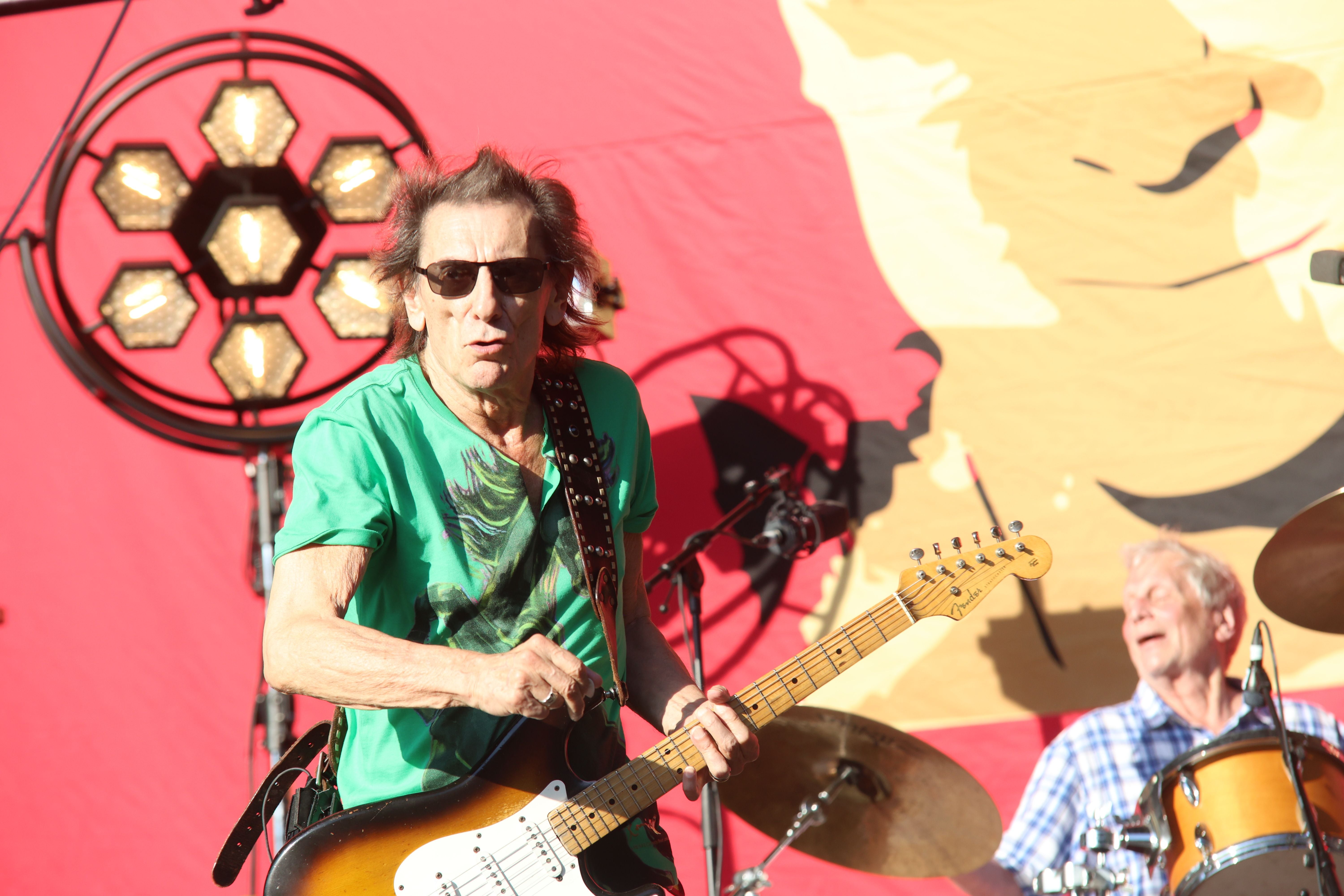 Gitarrist mit Sonnenbrille und grünem Shirt spielt auf einer Bühne, Schlagzeuger im Hintergrund.