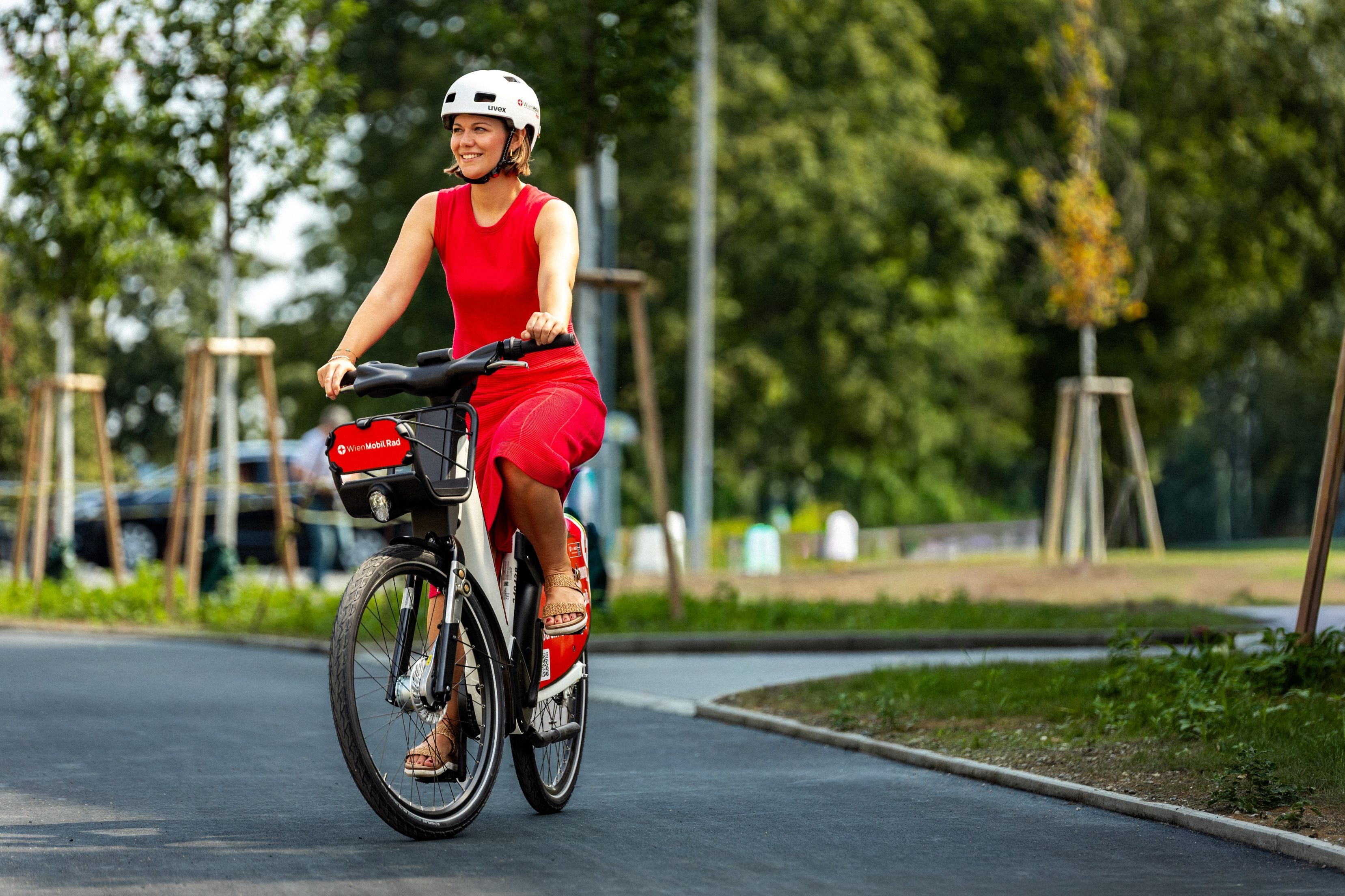 Frau mit rotem Kleid und Helm fährt ein E-Bike auf einem asphaltierten Weg im Grünen.