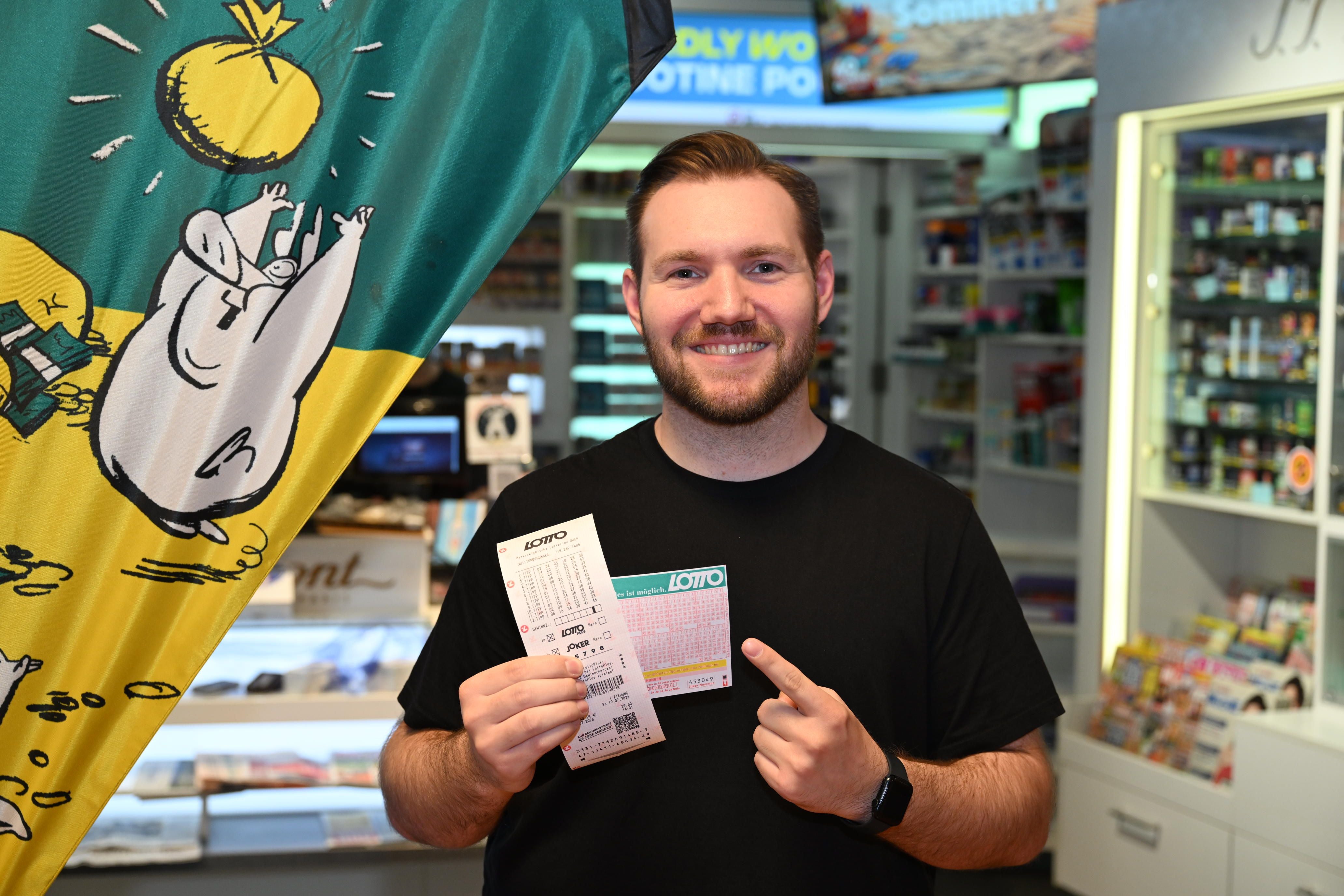 Ein lächelnder Mann hält zwei Lottoscheine in einem Kiosk und zeigt auf einen davon.