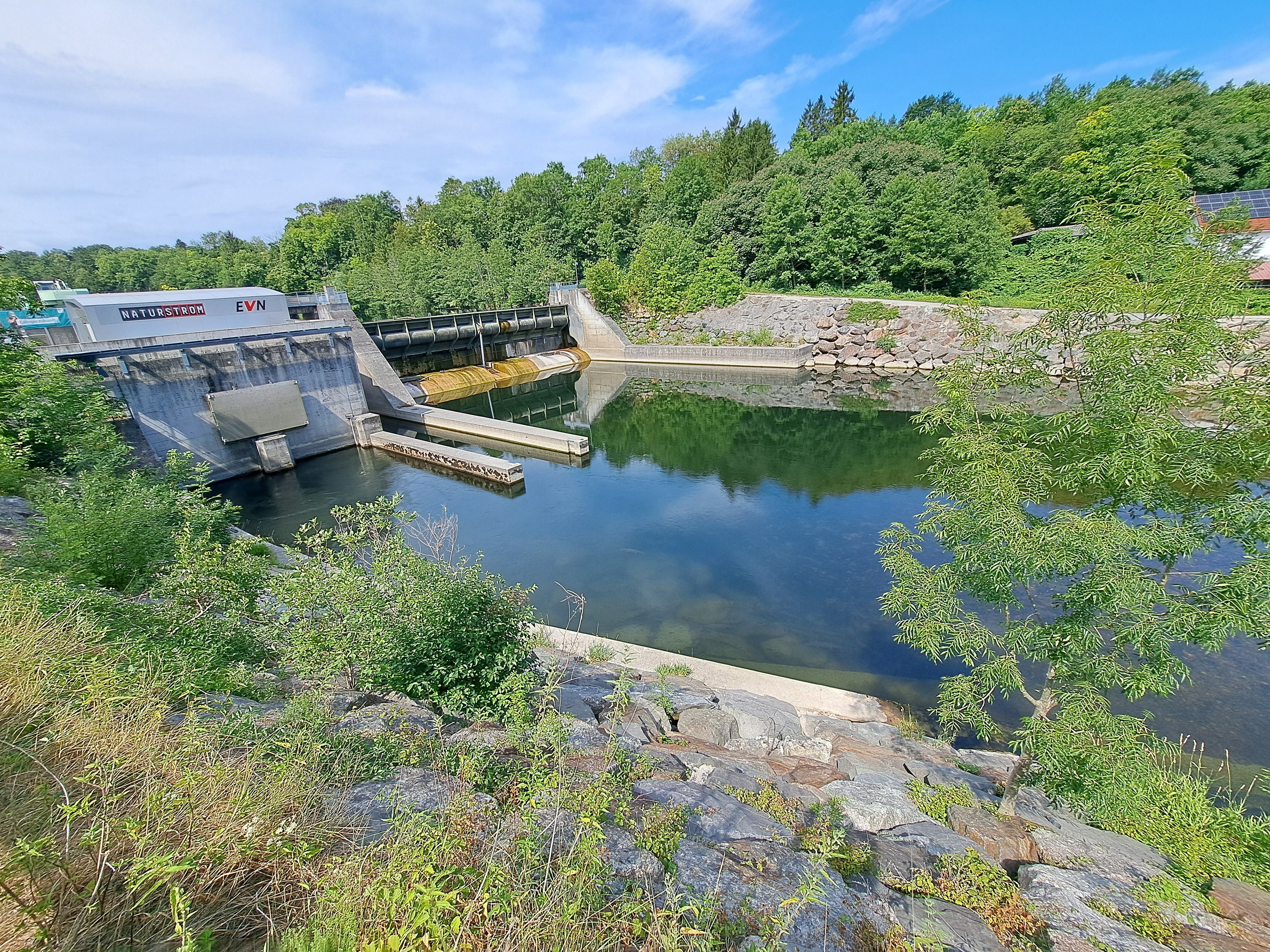 Kleines Wasserkraftwerk mit Stauwehr, umgeben von Bäumen und klarem Wasser.