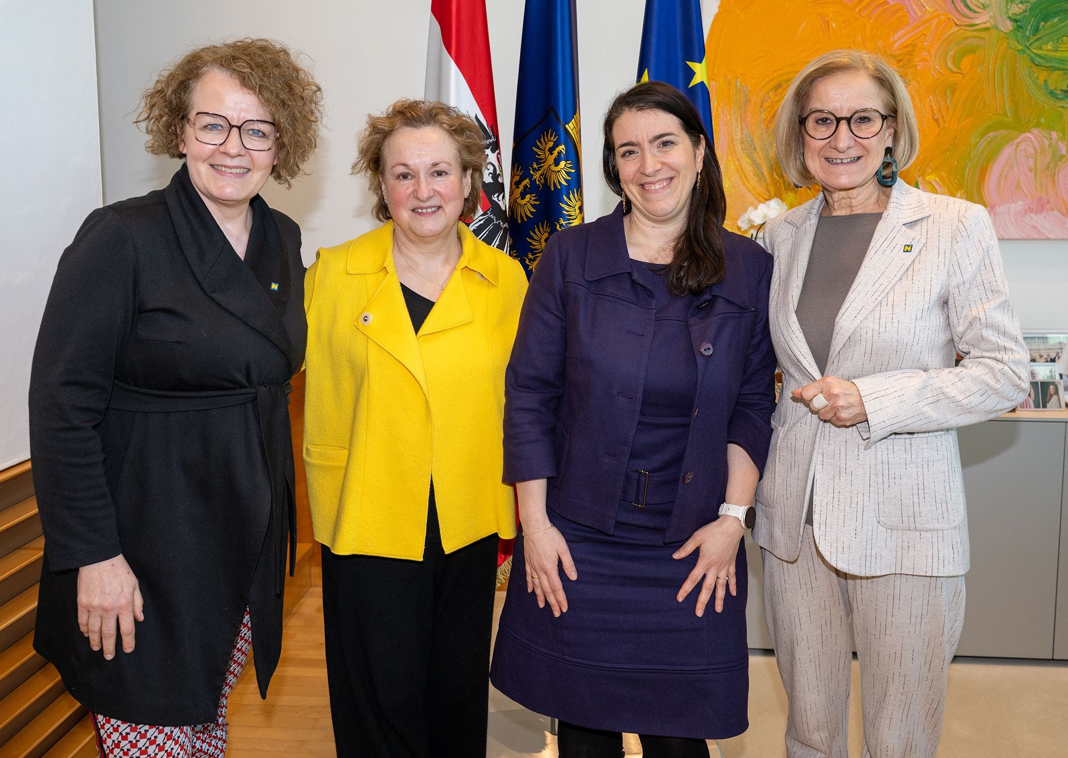 Vier Frauen posieren lächelnd vor österreichischer und EU-Flagge in einem Büro.
