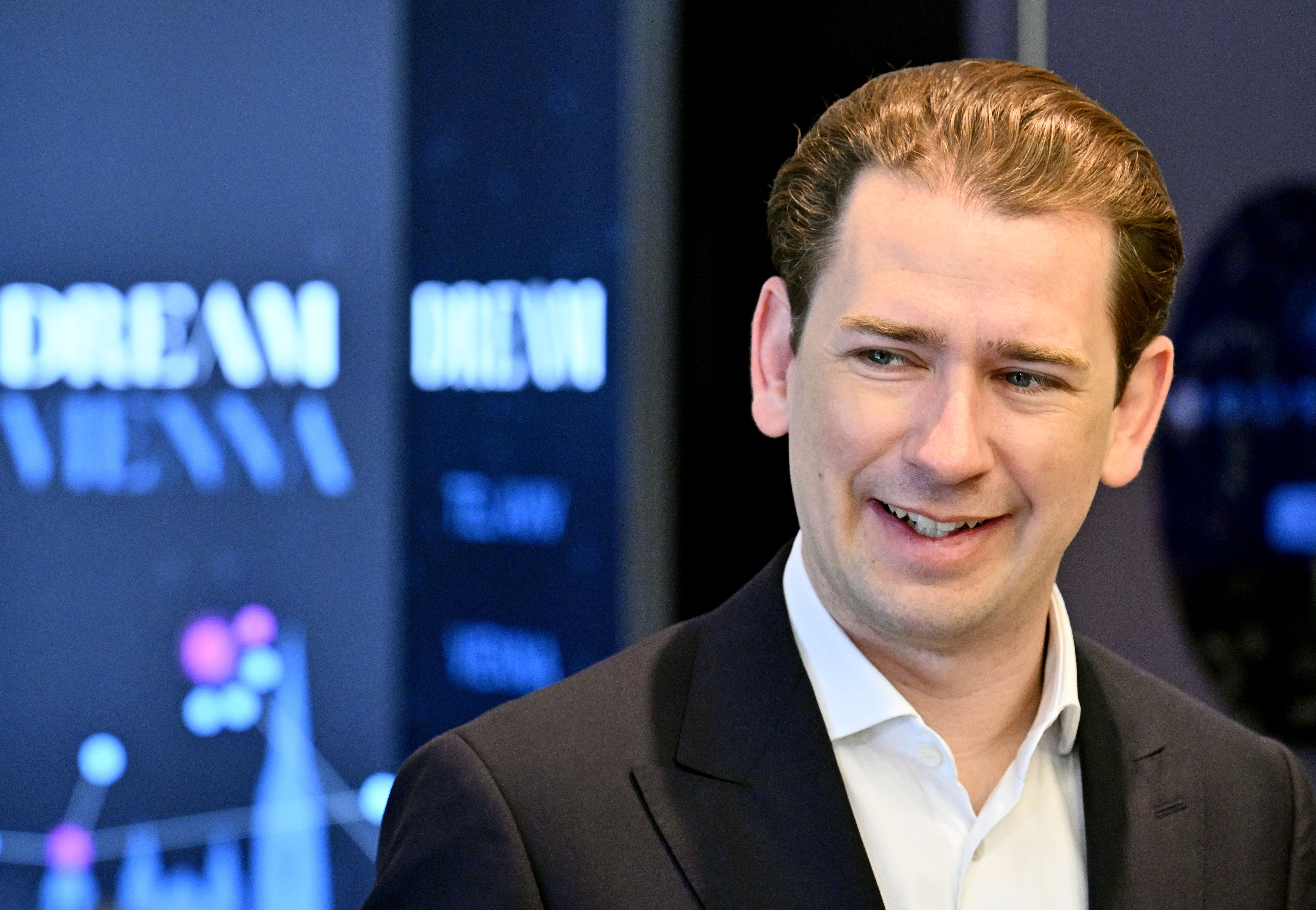 Ein Mann im Anzug lächelt bei einer Pressekonferenz mit unscharfem Hintergrund in Wien.
