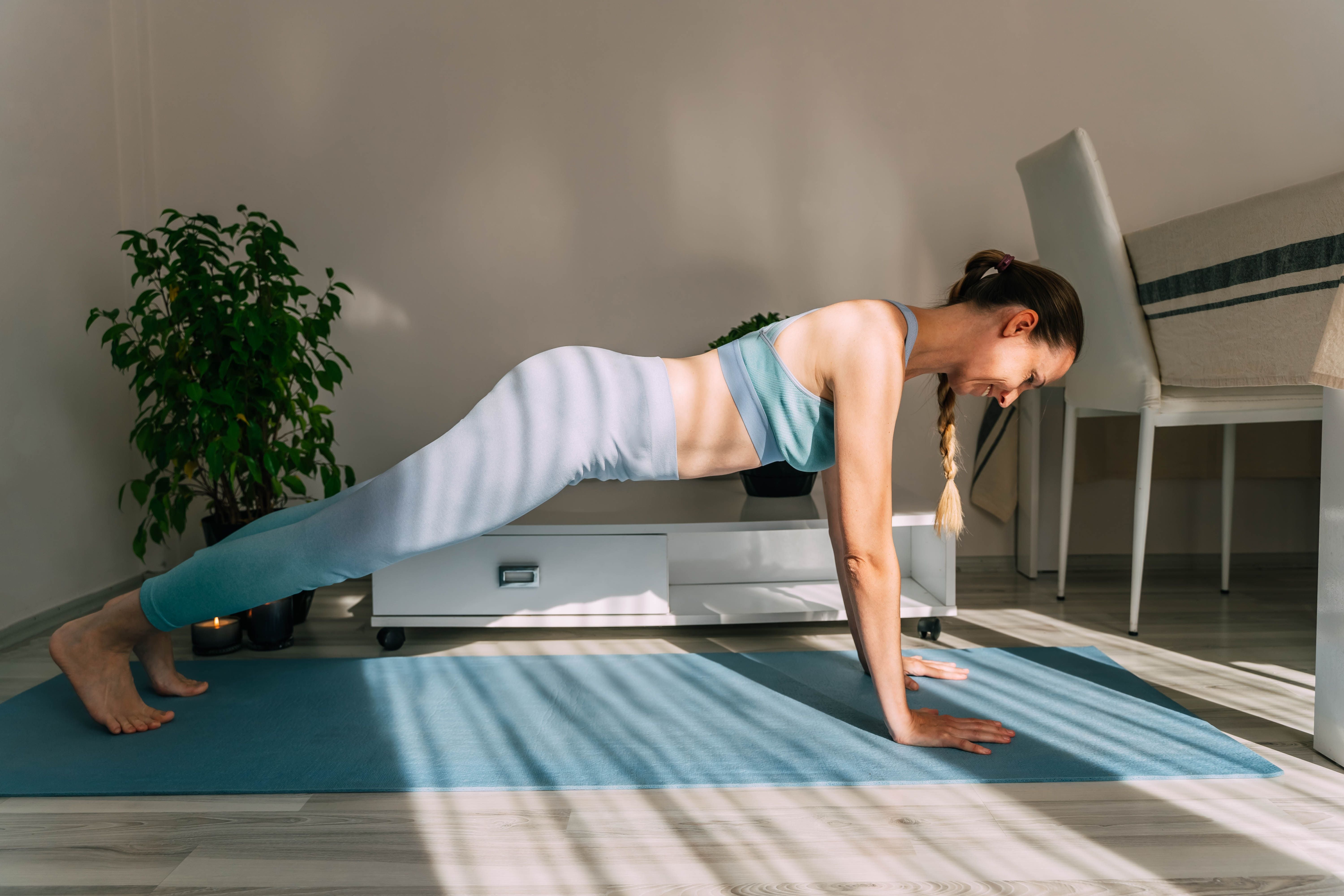Frau macht am sonnigen Tag eine Plank-Übung auf einer Yogamatte im Wohnzimmer.