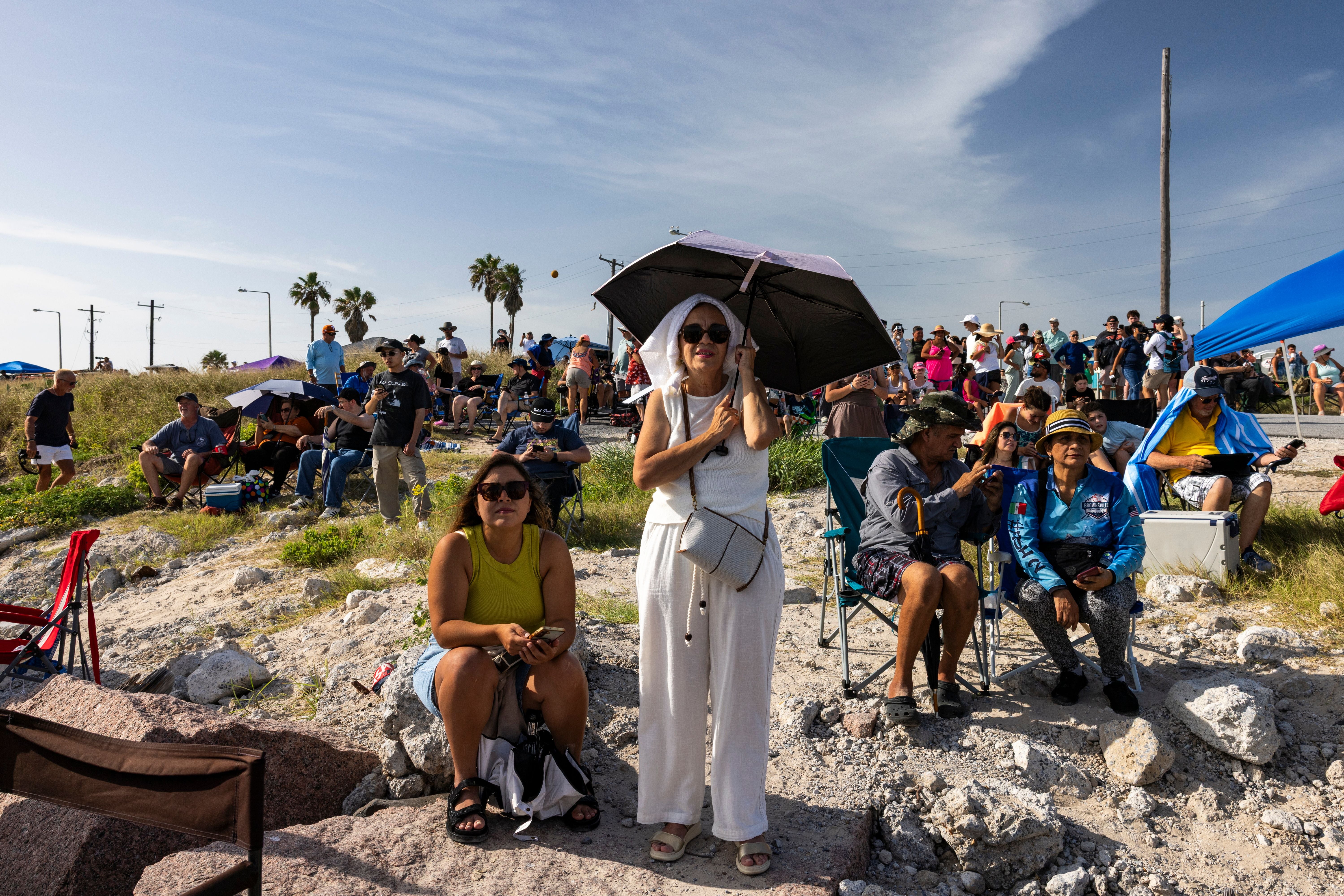 Zuschauer sitzen und stehen am Strand mit Sonnenschirmen, um einen Raketenstart zu beobachten.