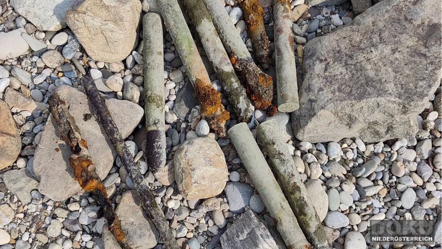 Mehrere rostige Munitionsreste liegen zwischen Steinen an einem Flussufer.