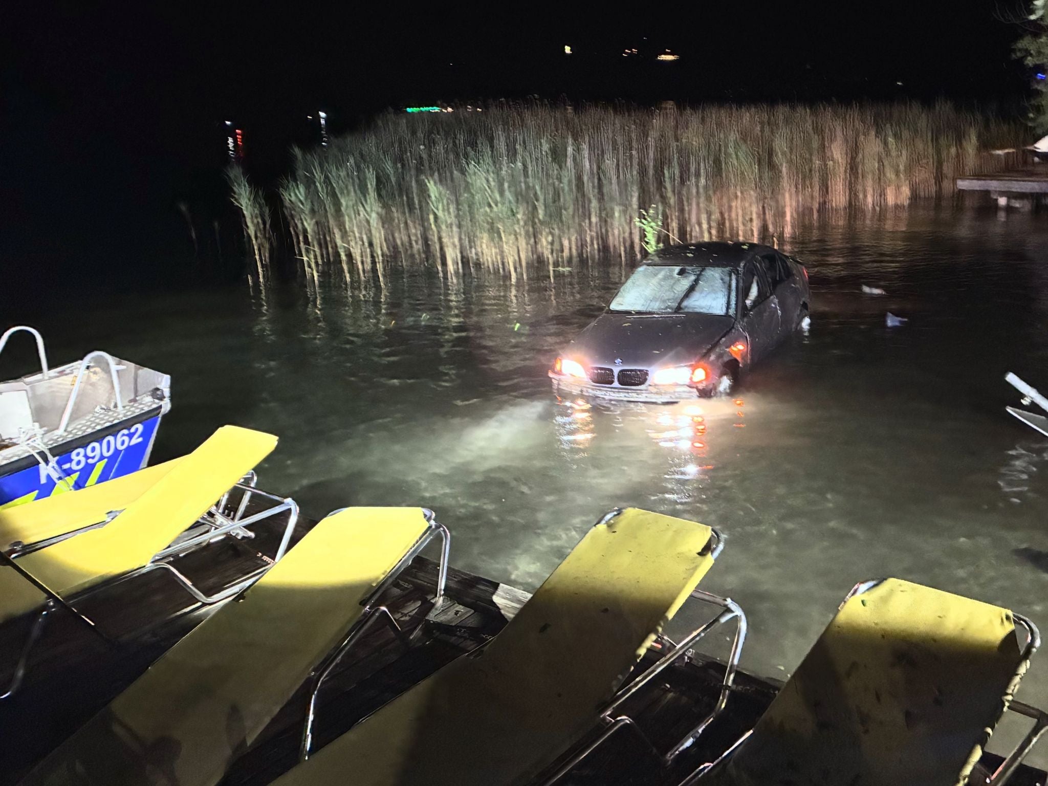 Ein Auto mit eingeschaltetem Licht steht nachts halb im Wasser an einem Seeufer, daneben Liegestühle.