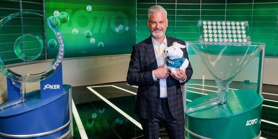 Ein Mann im Anzug hält ein Lotto-Maskottchen in einem Fernsehstudio für die Lottoziehung.