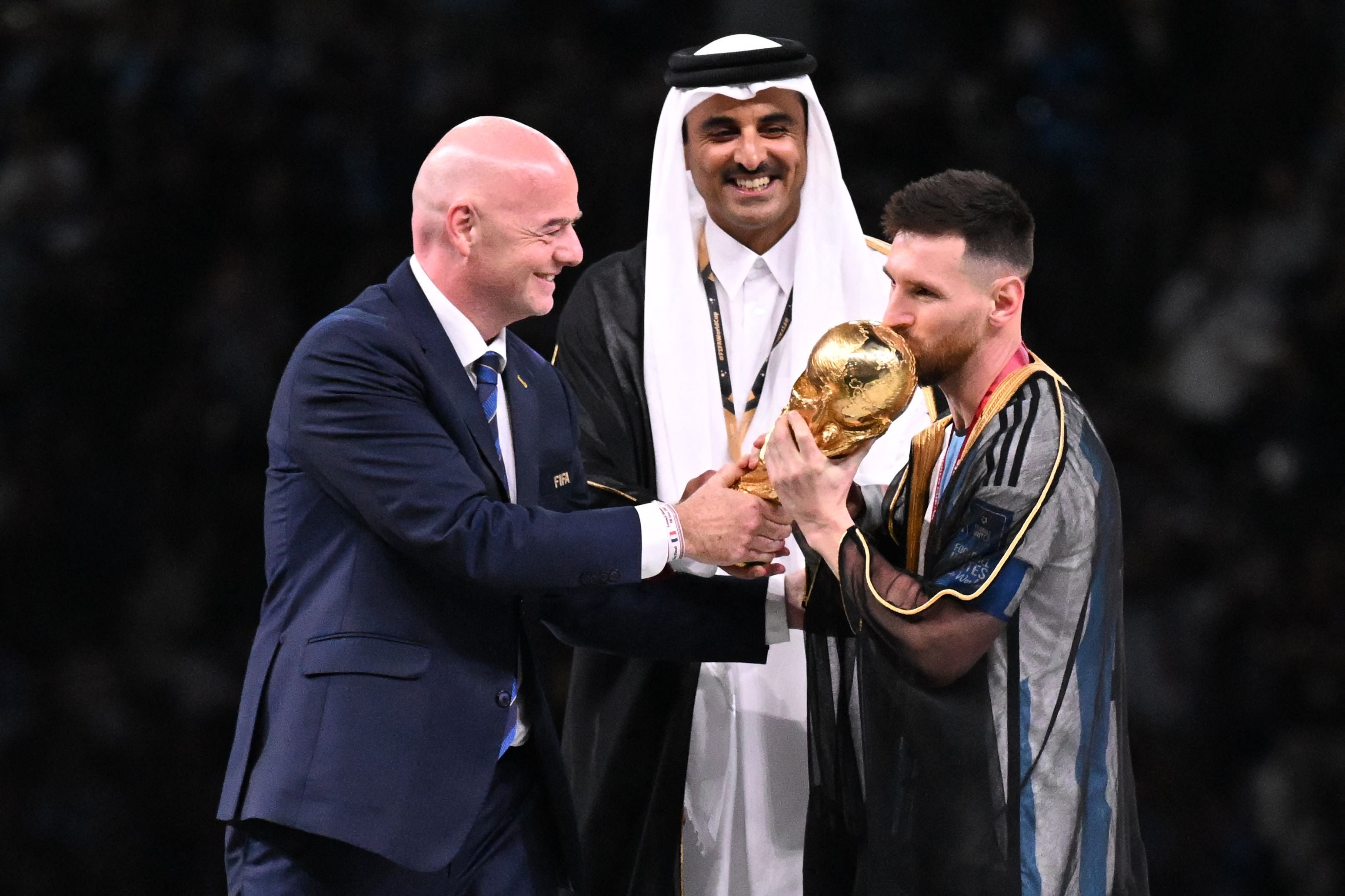 Lionel Messi küsst den WM-Pokal, daneben stehen Gianni Infantino und der Emir von Katar.