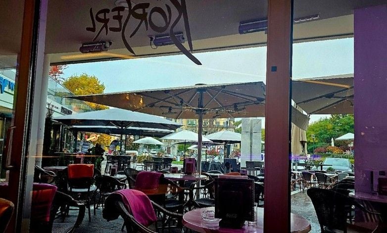Blick von innen auf ein Café mit leeren Stühlen, Tischen und großen Sonnenschirmen draußen.