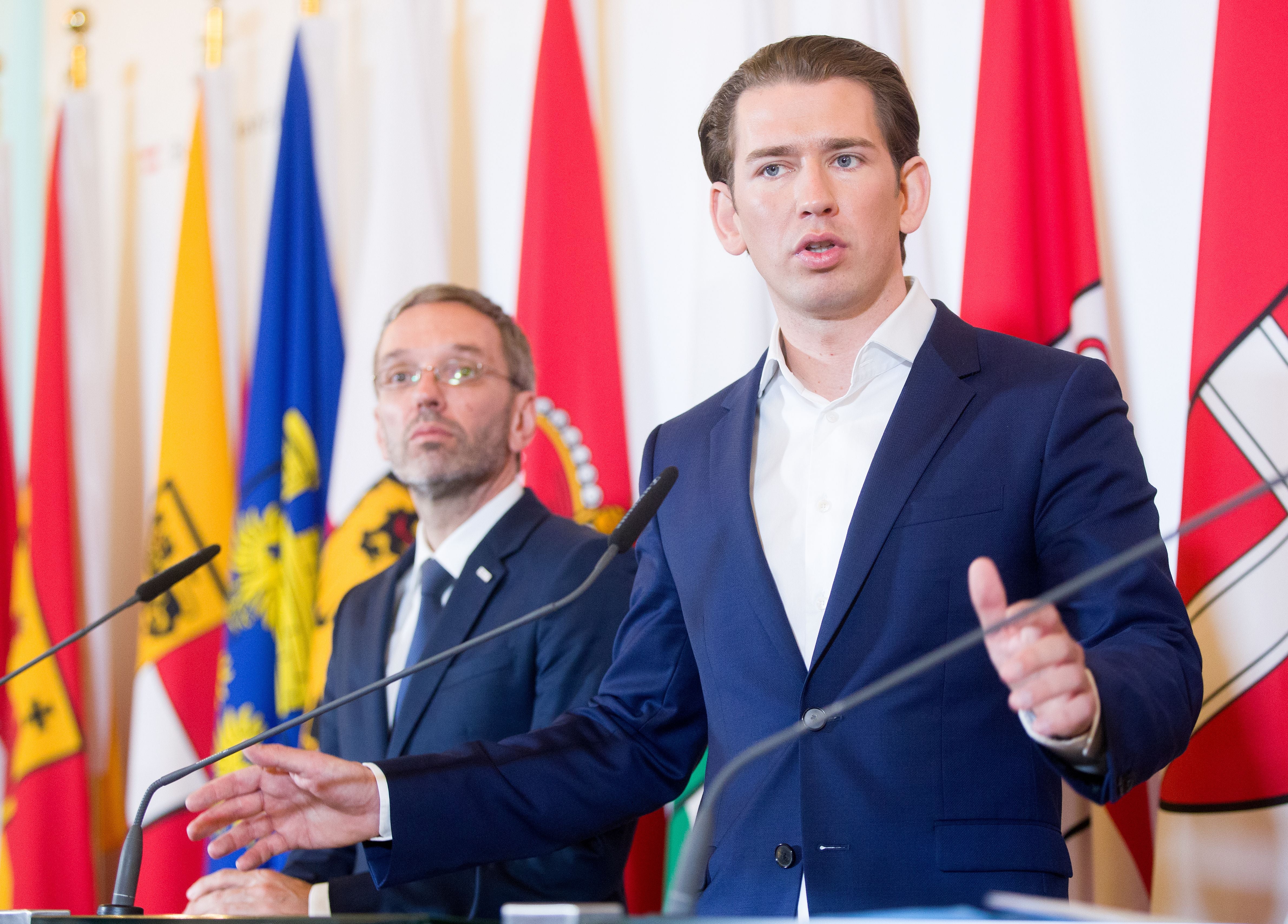 Zwei Männer im Anzug sprechen bei einer Pressekonferenz vor mehreren Länderflaggen.