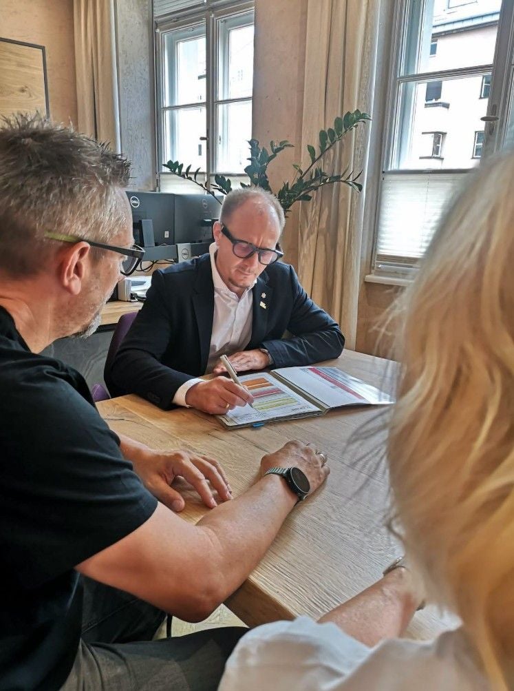 Drei Personen sitzen an einem Tisch, einer erklärt etwas in einem Ordner mit bunten Tabellen.