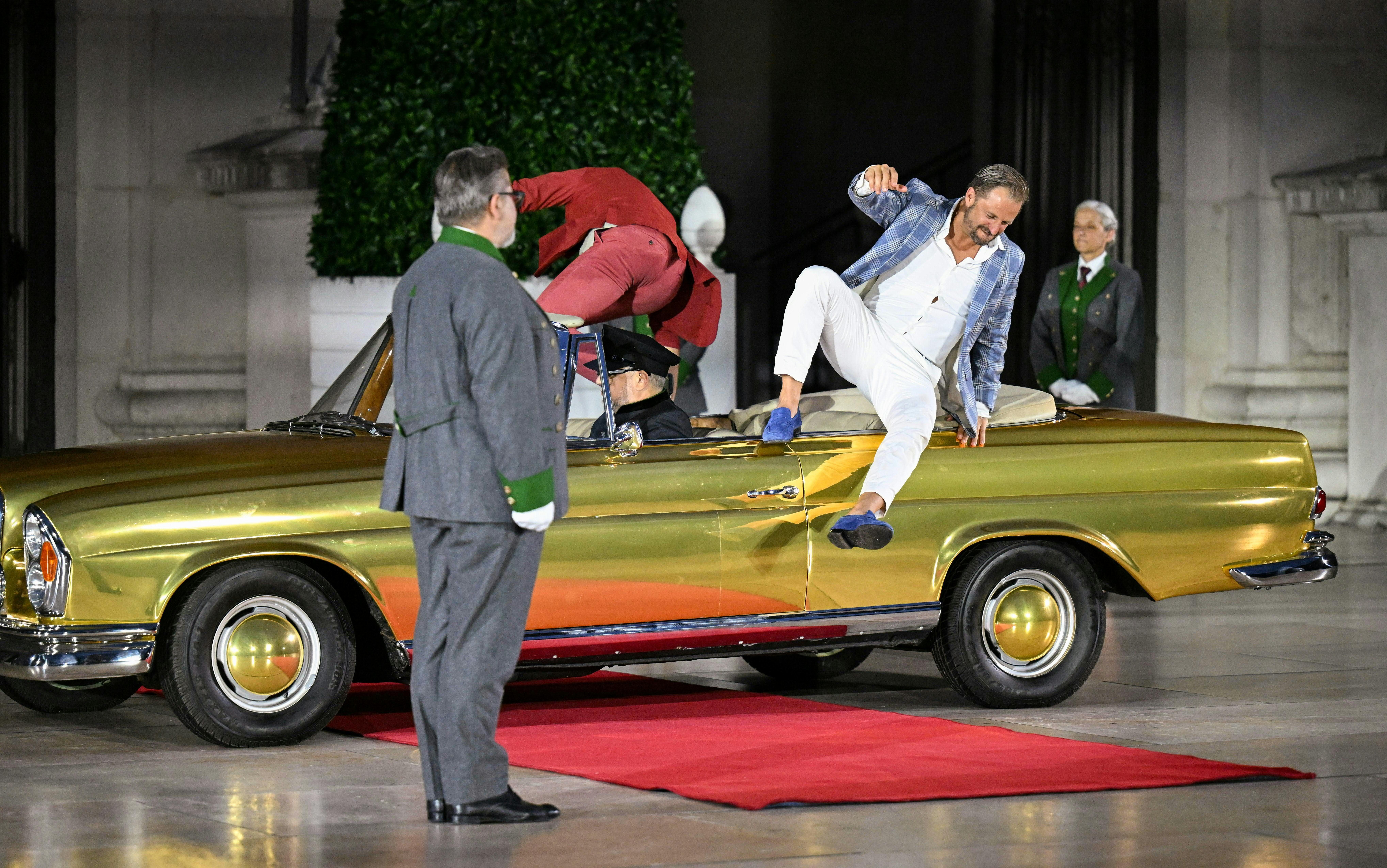 Zwei Männer steigen bei einer Theateraufführung aus einem goldenen Auto auf einem roten Teppich.