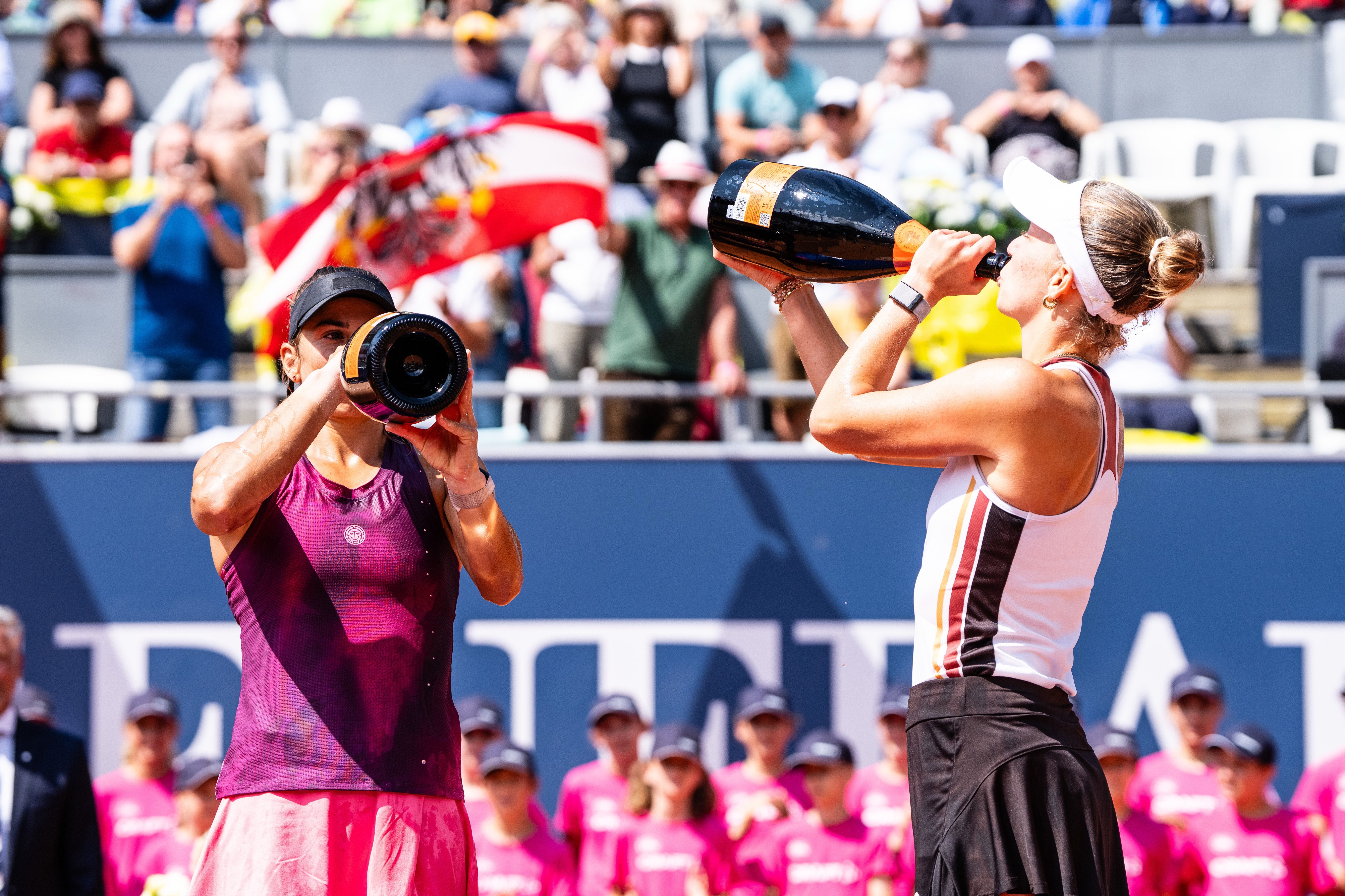 Zwei Tennisspielerinnen trinken mit Sektflaschen auf dem Tennisplatz, Zuschauer im Hintergrund.
