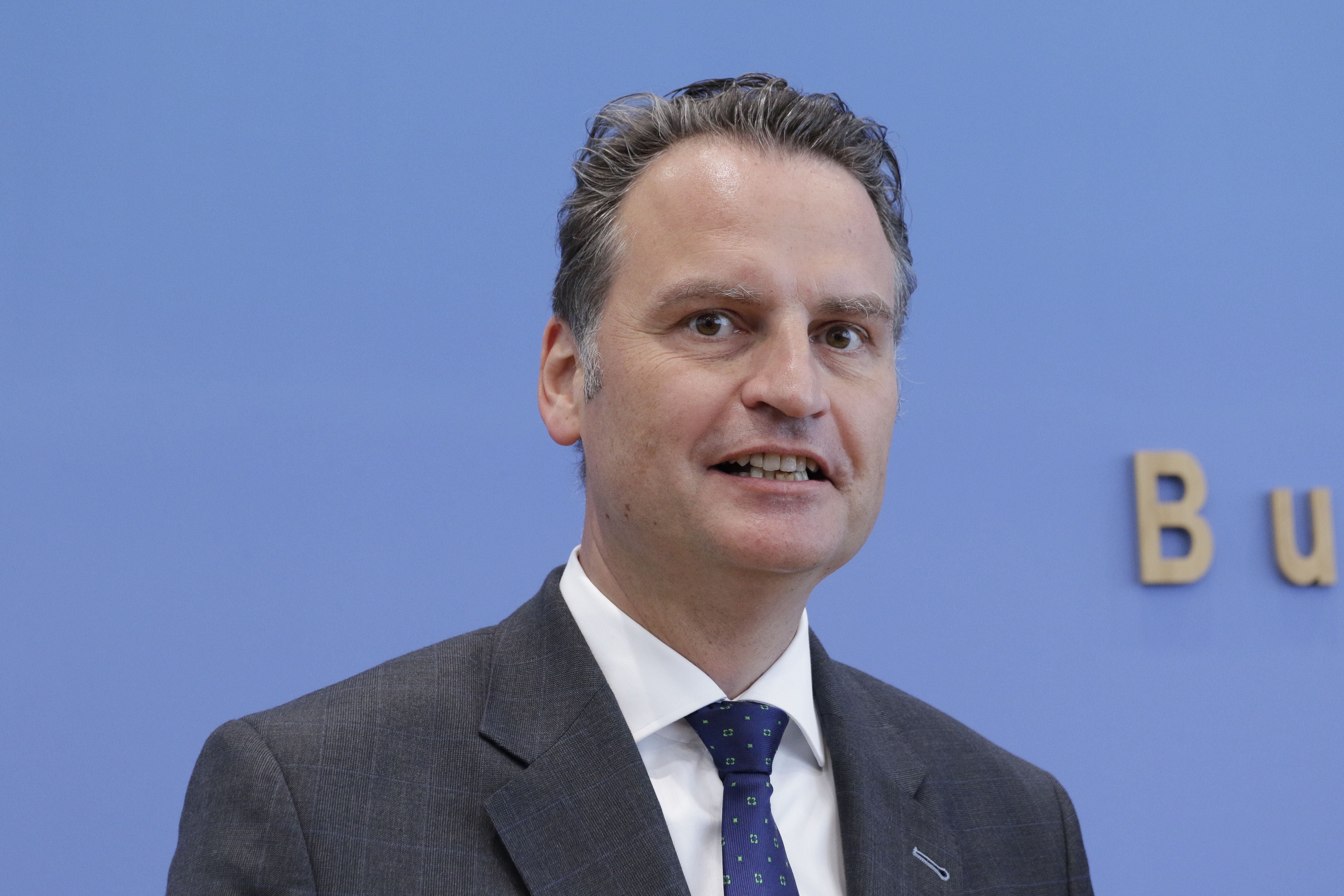 Ein Mann im Anzug spricht vor blauem Hintergrund mit einem Teil eines Schriftzugs im Bild.