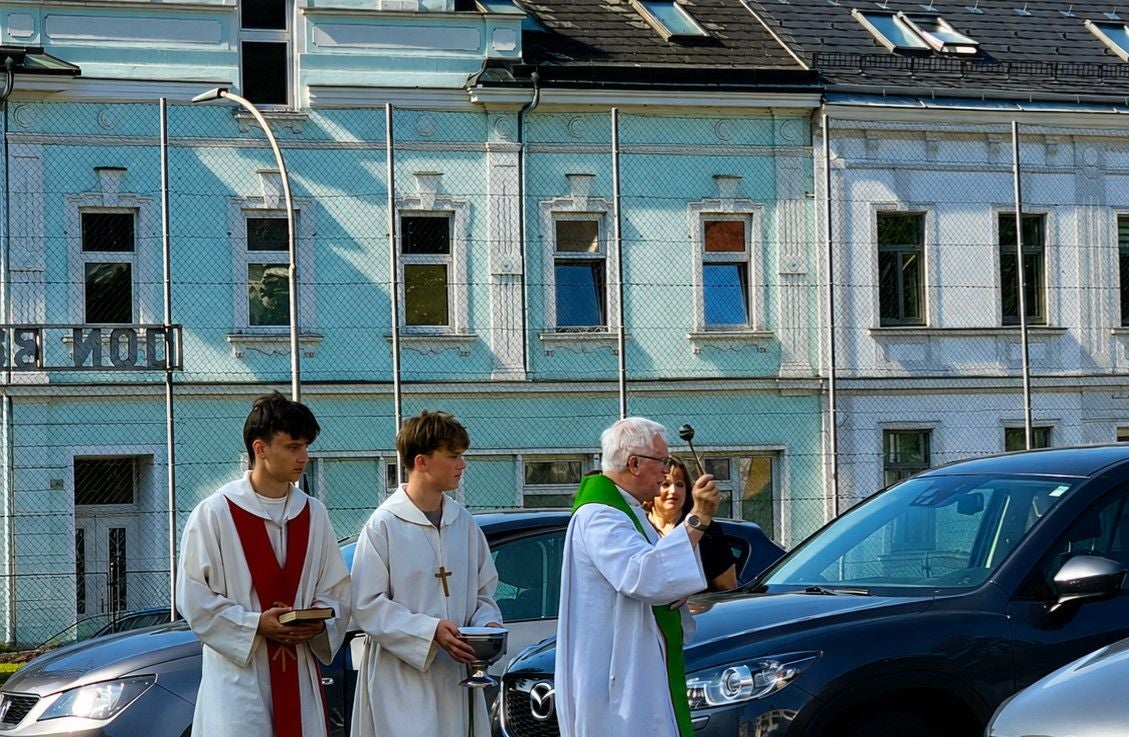 Ein Priester segnet Autos im Freien, begleitet von zwei Ministranten in liturgischer Kleidung.
