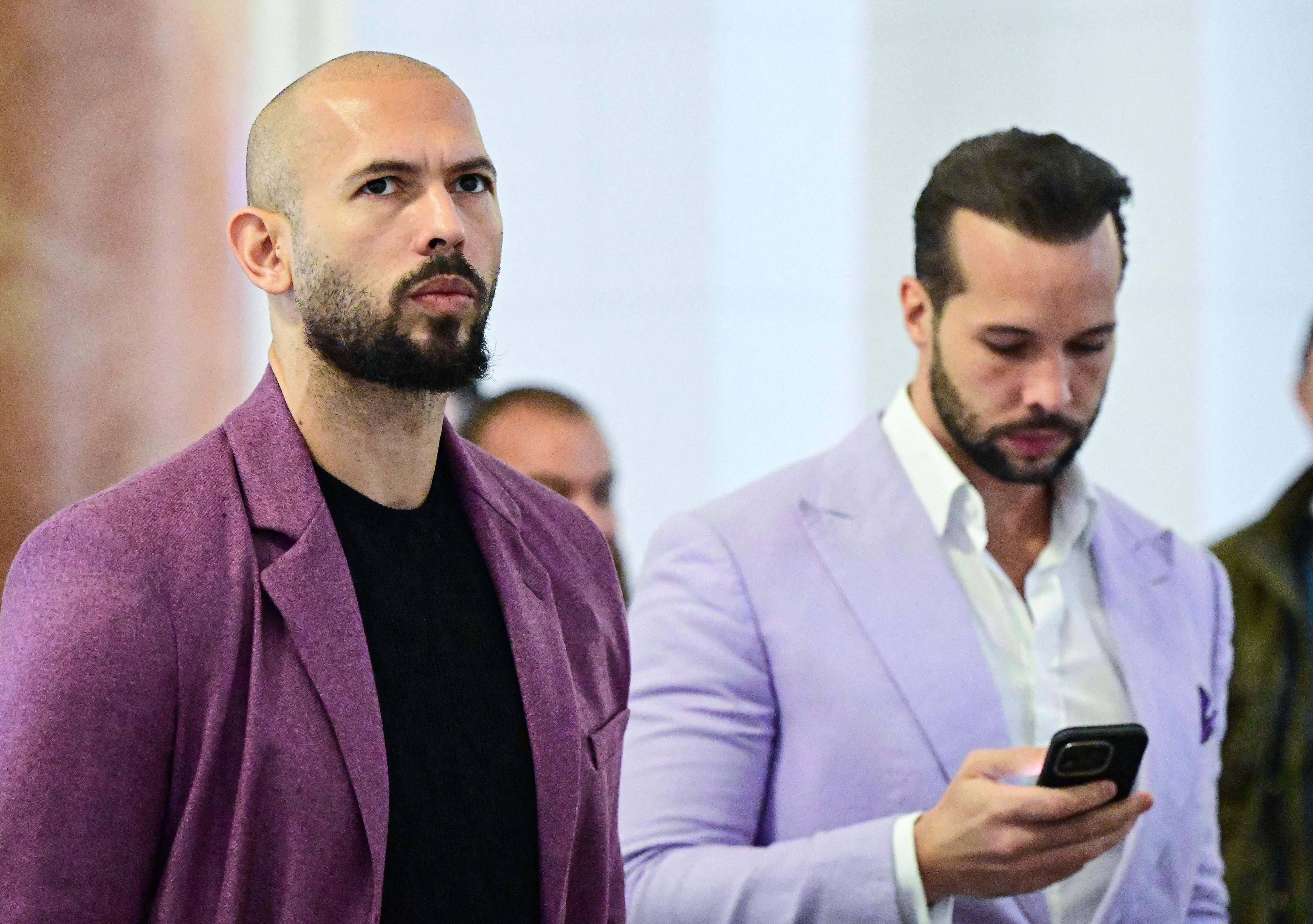 Zwei Männer in lila Sakkos stehen nebeneinander, einer schaut ernst, der andere nutzt ein Smartphone.