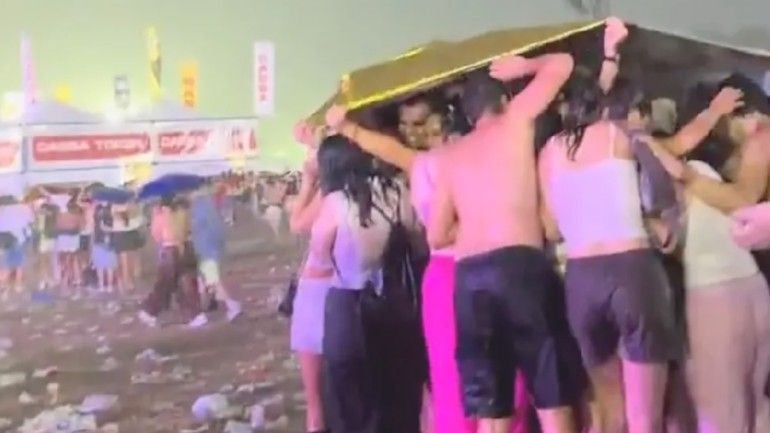 Menschenmenge sucht Schutz vor Regen unter einer Plane auf einem nassen Festivalgelände.