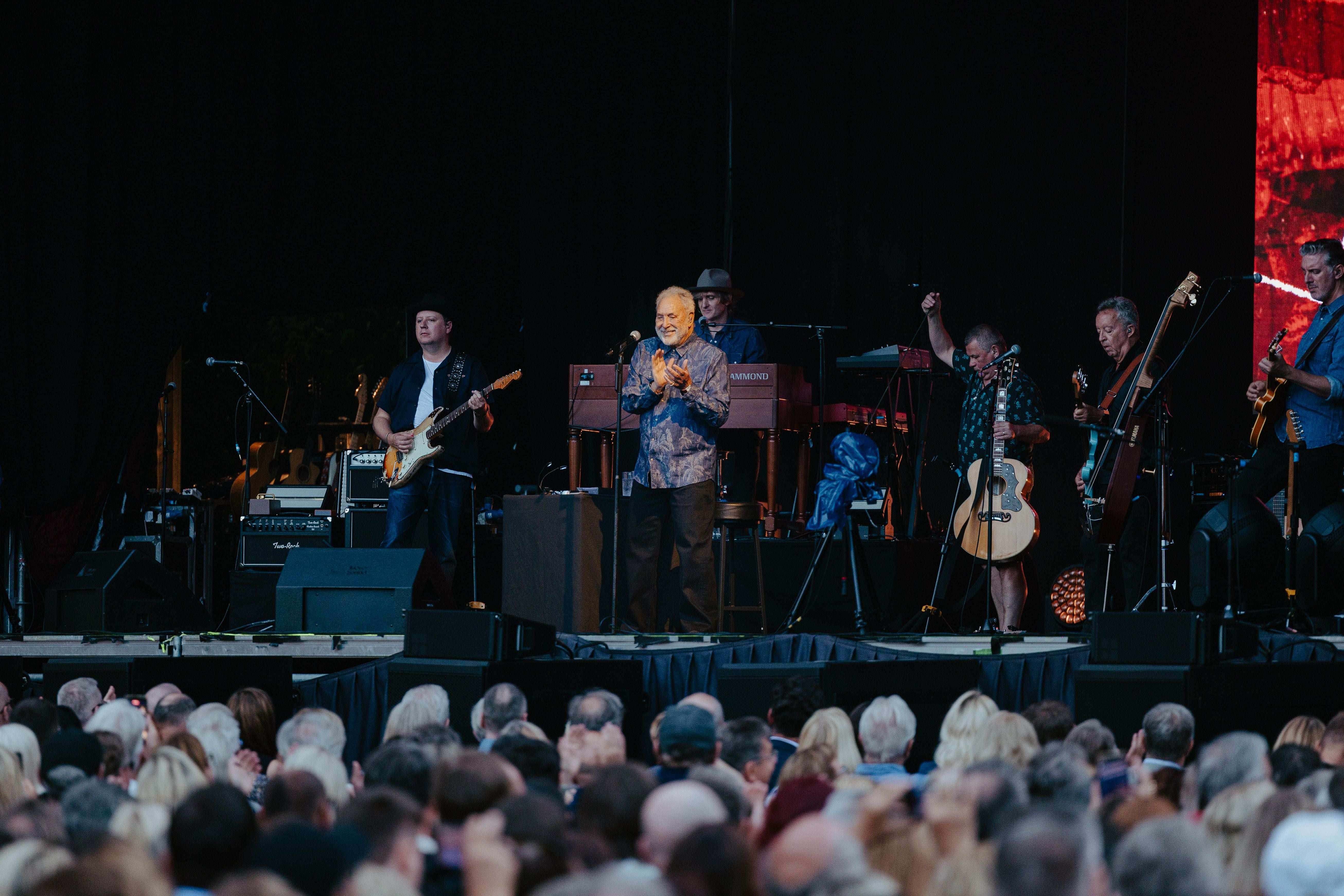 Tom Jones und seine Band treten auf einer Konzertbühne vor großem Publikum auf.