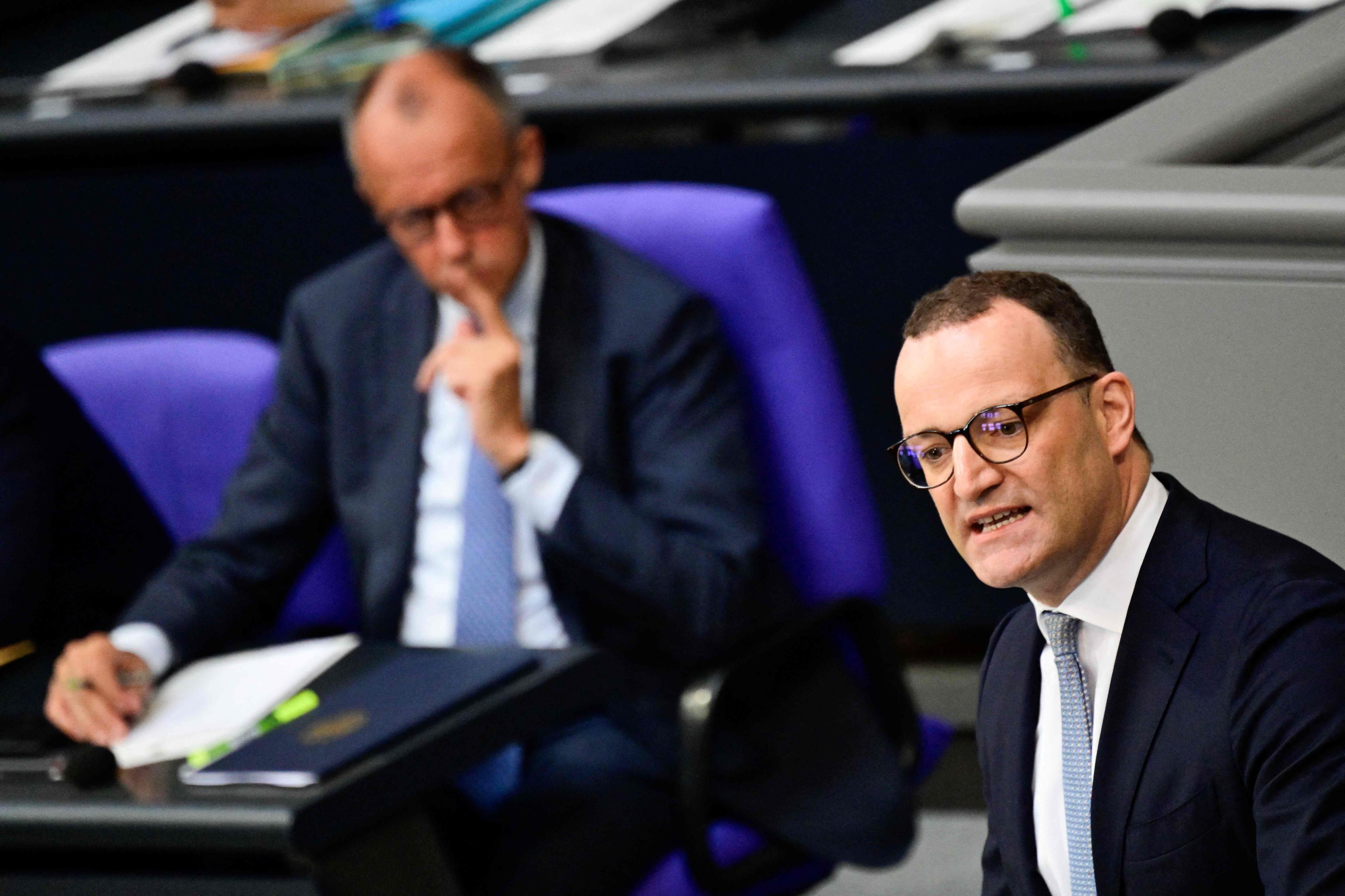 Jens Spahn spricht im Bundestag, Friedrich Merz sitzt unscharf im Hintergrund auf lila Stuhl.