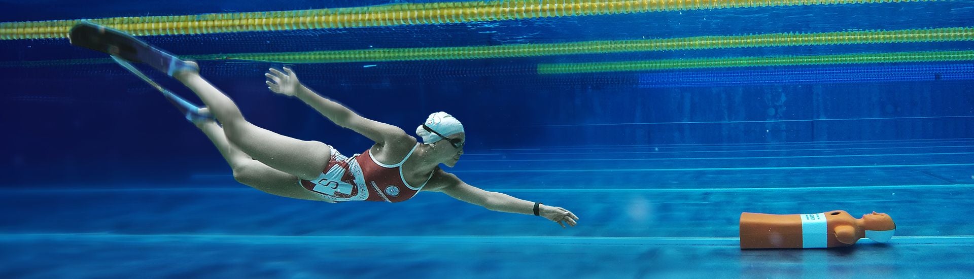 Taucherin schwimmt unter Wasser auf eine Rettungspuppe im Schwimmbecken zu.