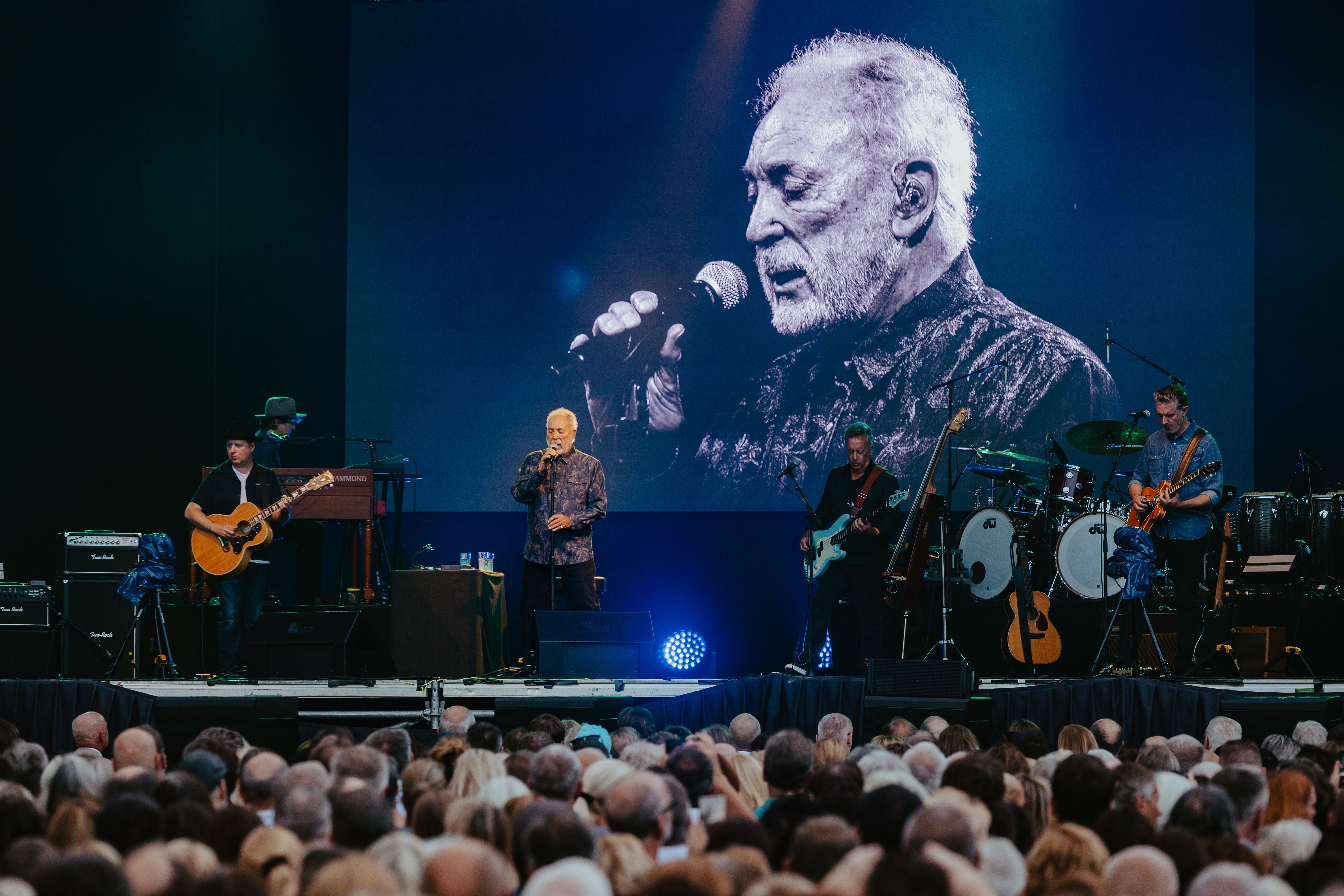Tom Jones singt mit Band bei einem Live-Konzert, große Leinwand zeigt sein Porträt.