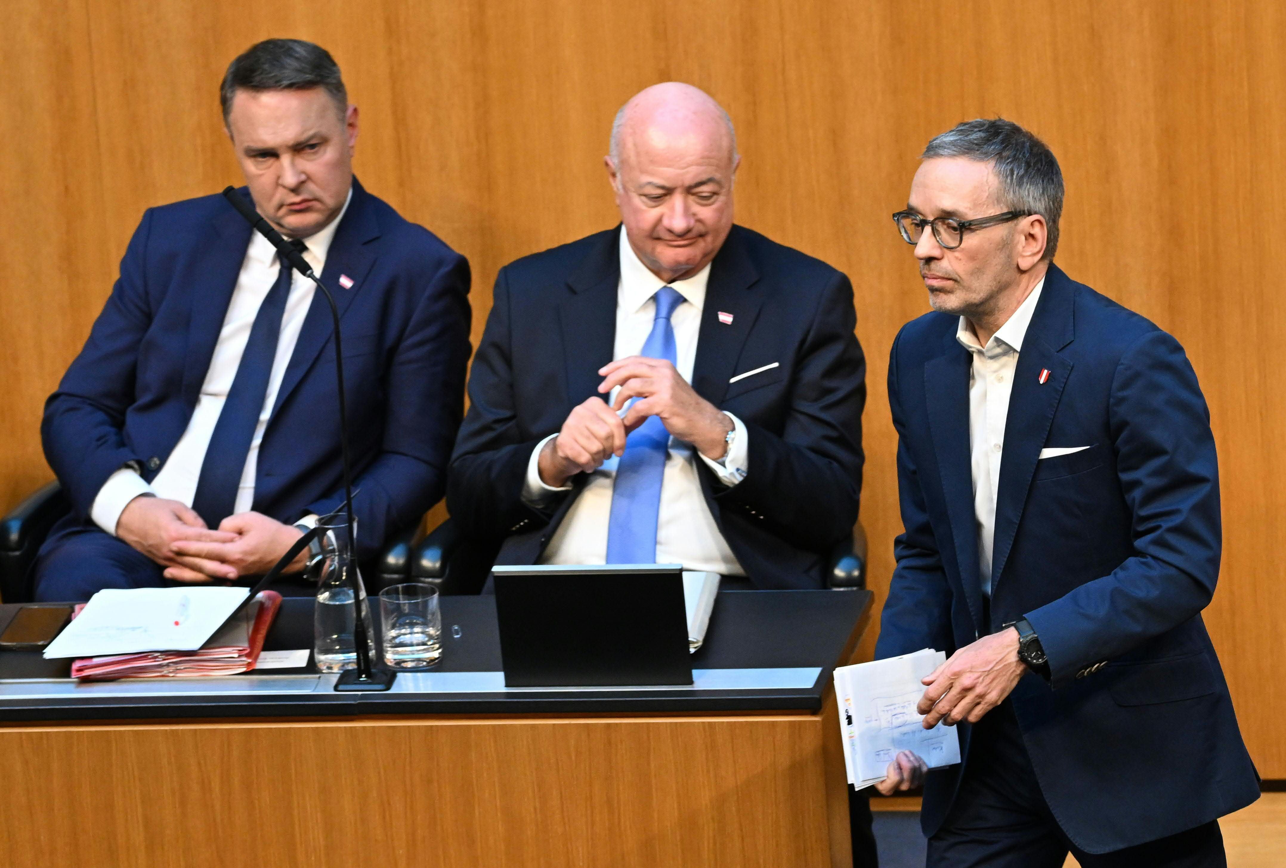 Drei Männer im Anzug bei einer Parlamentssitzung in Wien, einer steht, zwei sitzen.