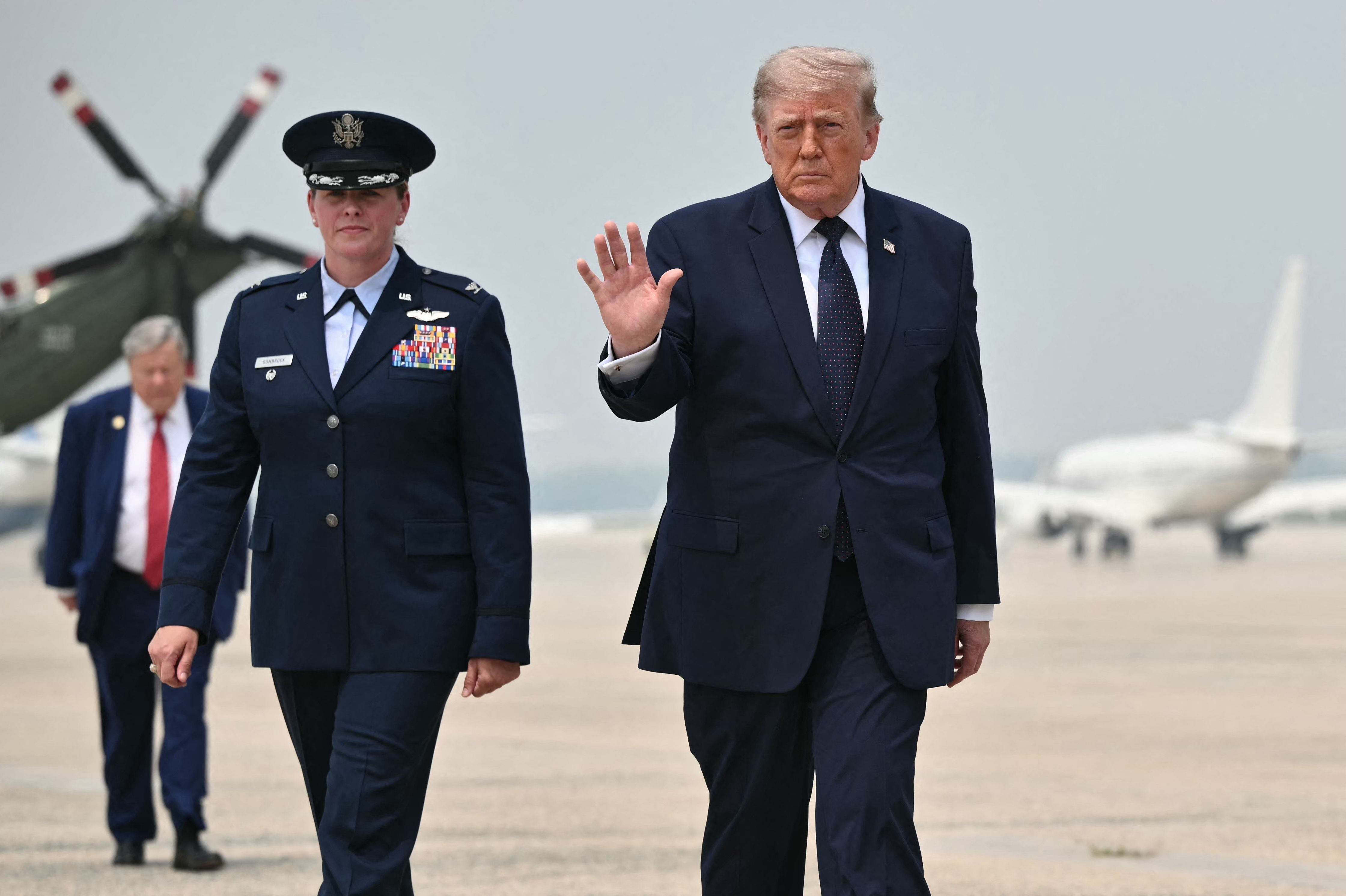 Donald Trump winkt am Flughafen, begleitet von einer Militärangehörigen, im Hintergrund Flugzeuge.