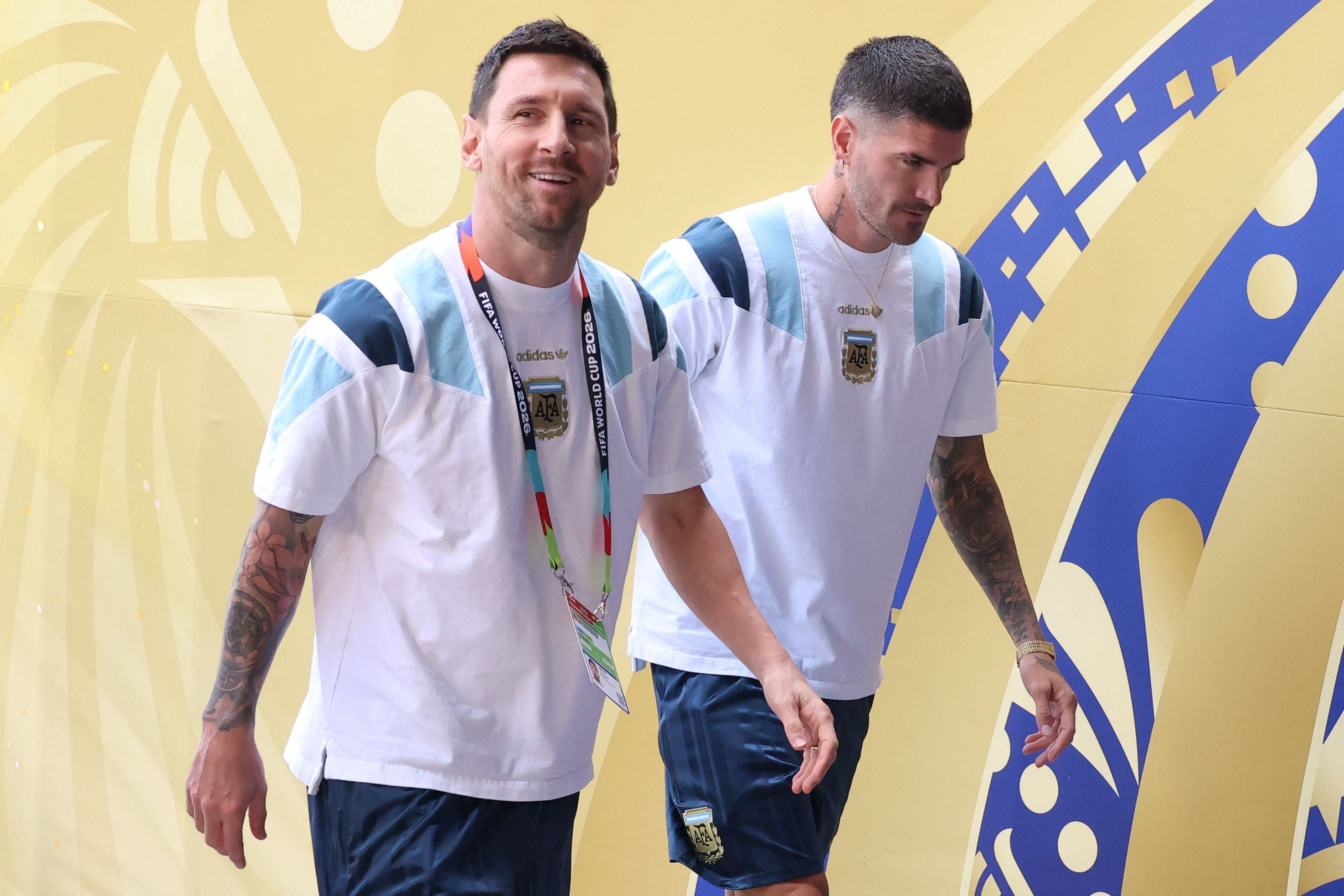 Zwei argentinische Fußballspieler betreten gemeinsam ein Stadion mit gelbem Hintergrund.