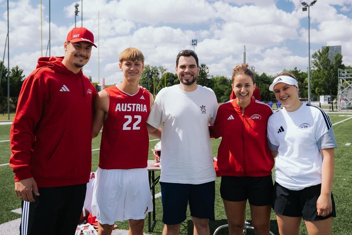 Fünf junge Leute, teils in Österreich-Sportkleidung, stehen lachend auf einem Sportplatz.